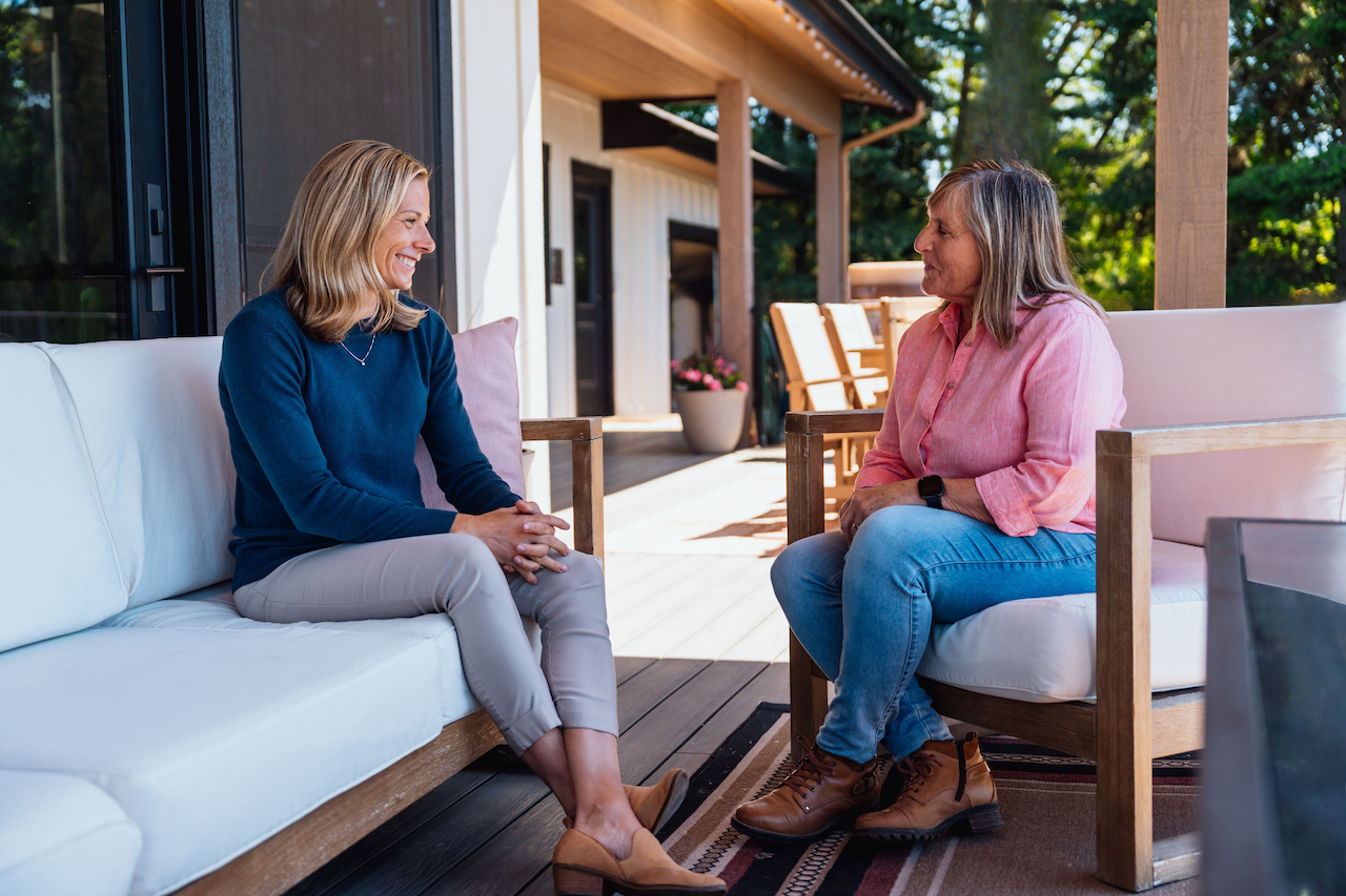 Two women sitting on outdoor furniture, talking and smiling, on a porch with trees in the background.