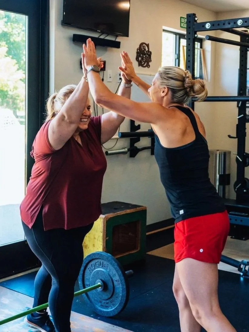 Two women celebrating with a high-five in a gym, one woman wearing a maroon shirt and black leggings, the other woman wearing a black tank top and red shorts, weightlifting equipment in the background.
