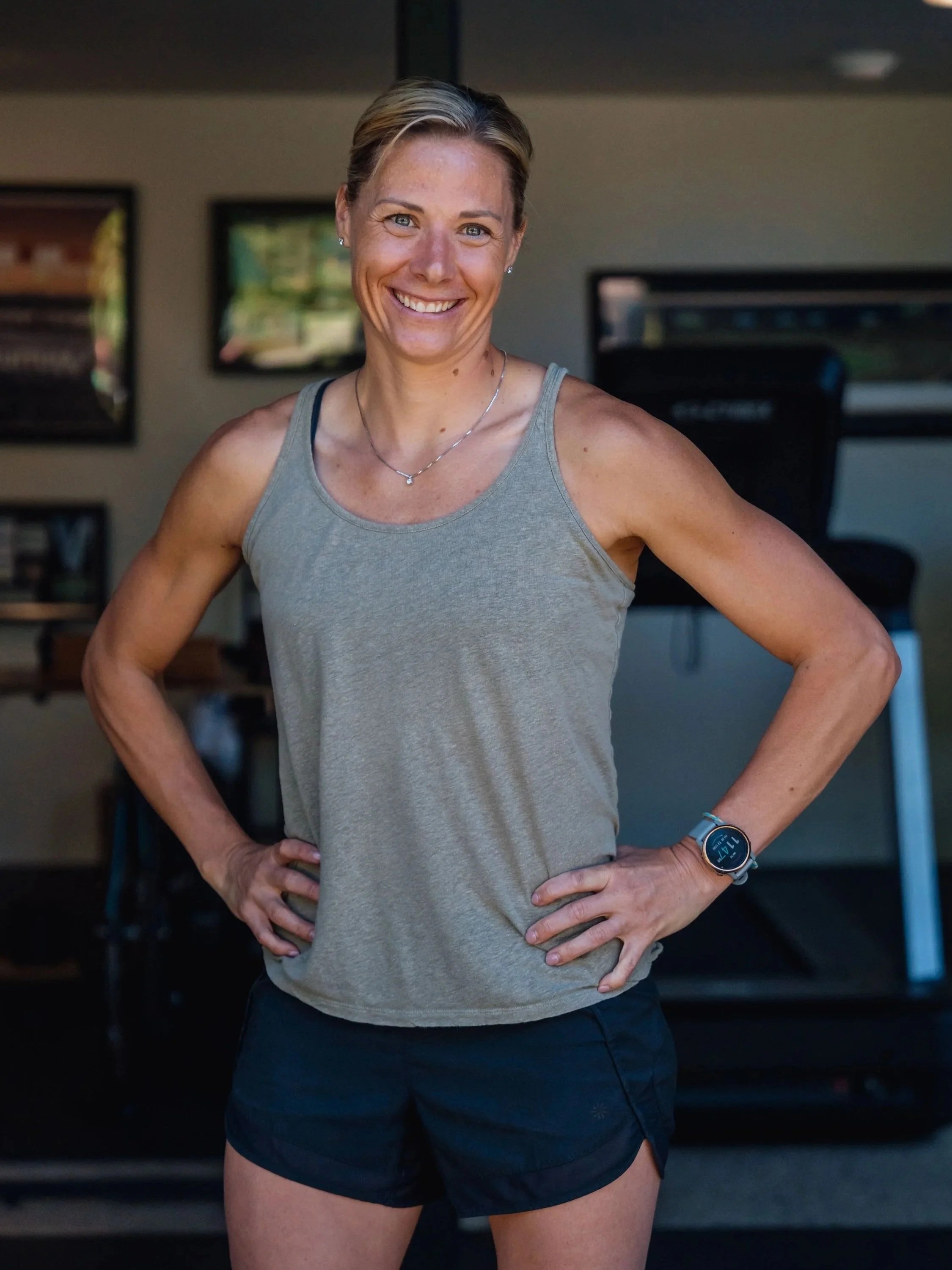 A smiling woman with her hands on her hips wearing a gray tank top, black shorts, a silver watch, and a necklace, standing indoors in front of exercise equipment.