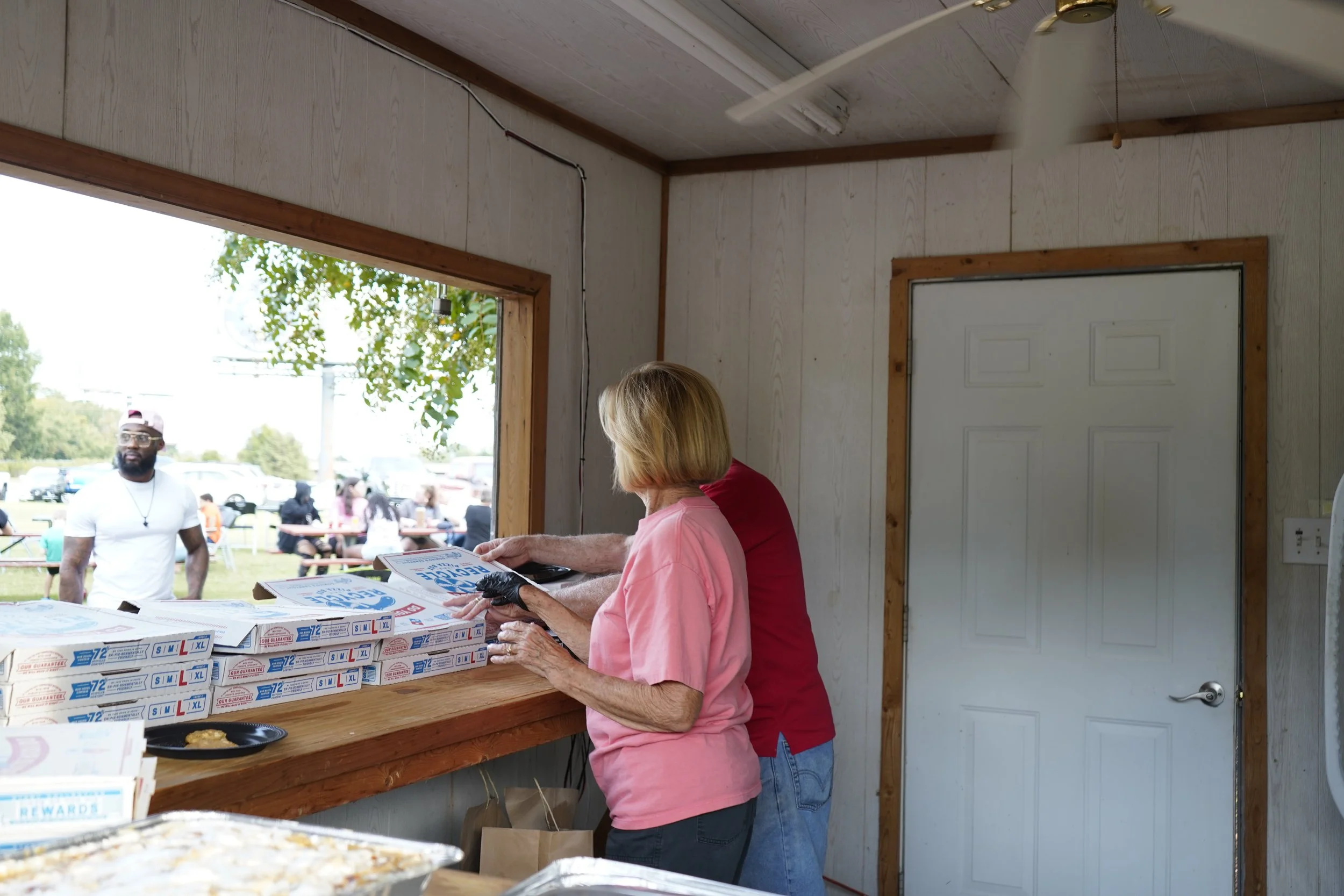 An elderly woman and a man looking at food orders through a counter window at an outdoor event, with pizza boxes and paper bags on the counter, and people in the background.