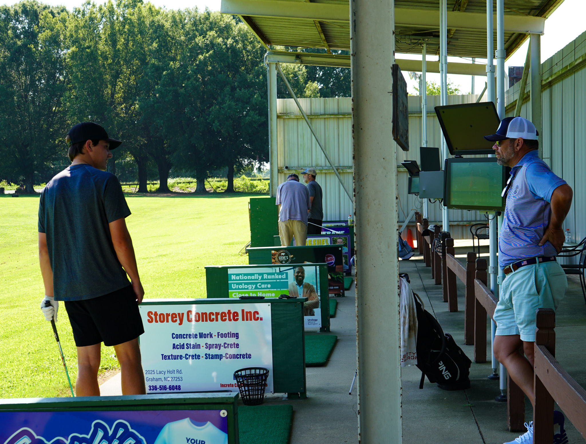 People at a golf driving range. A young man in a dark shirt and cap holding a golf club stands near a sign advertising concrete work services. Two older men are near the shooting range, one with a golf bag and the other looking at multiple screens. Green grass and trees are in the background.