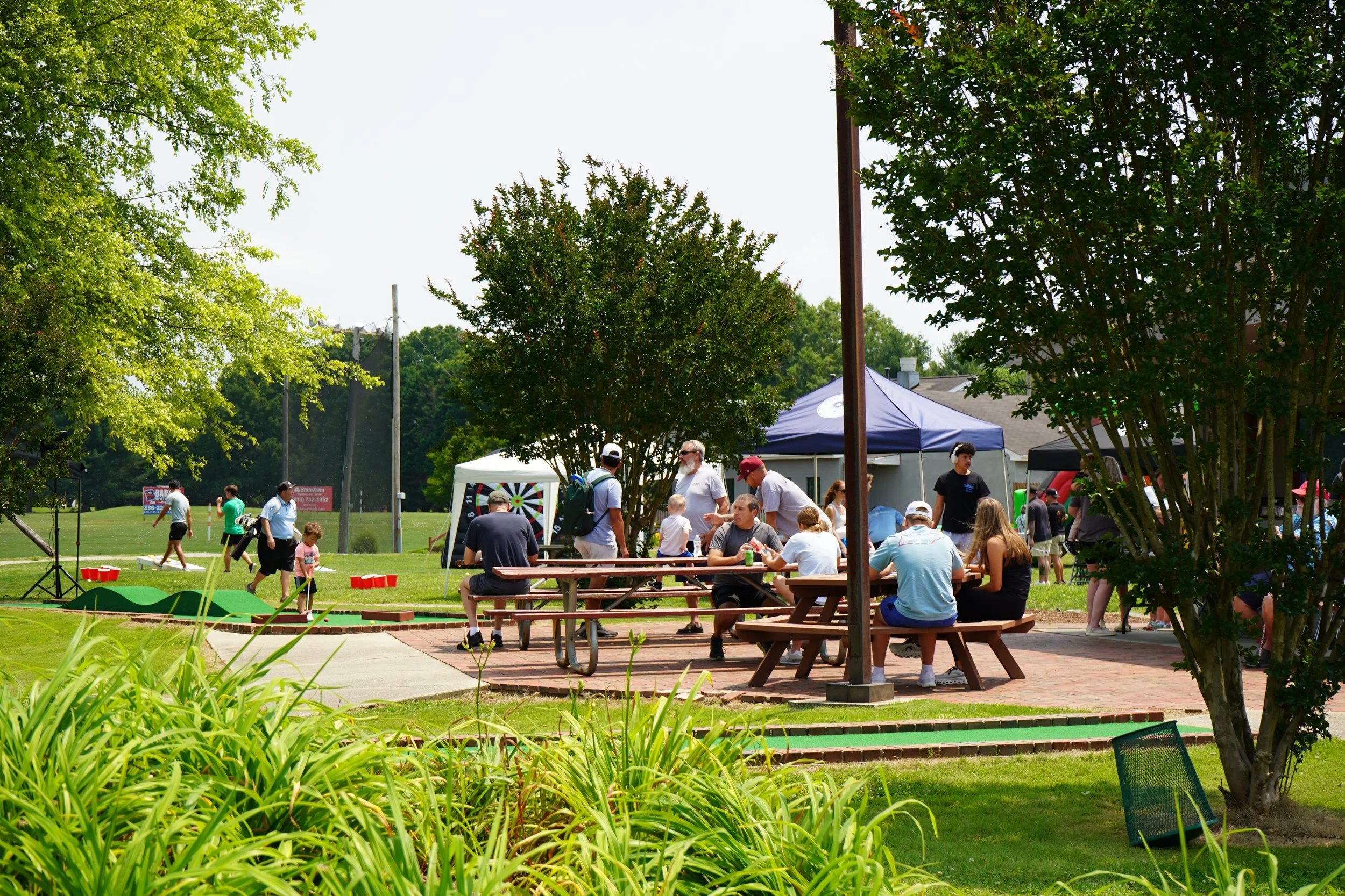 People gathered at an outdoor park event with picnic tables and tents, surrounded by trees and greenery.
