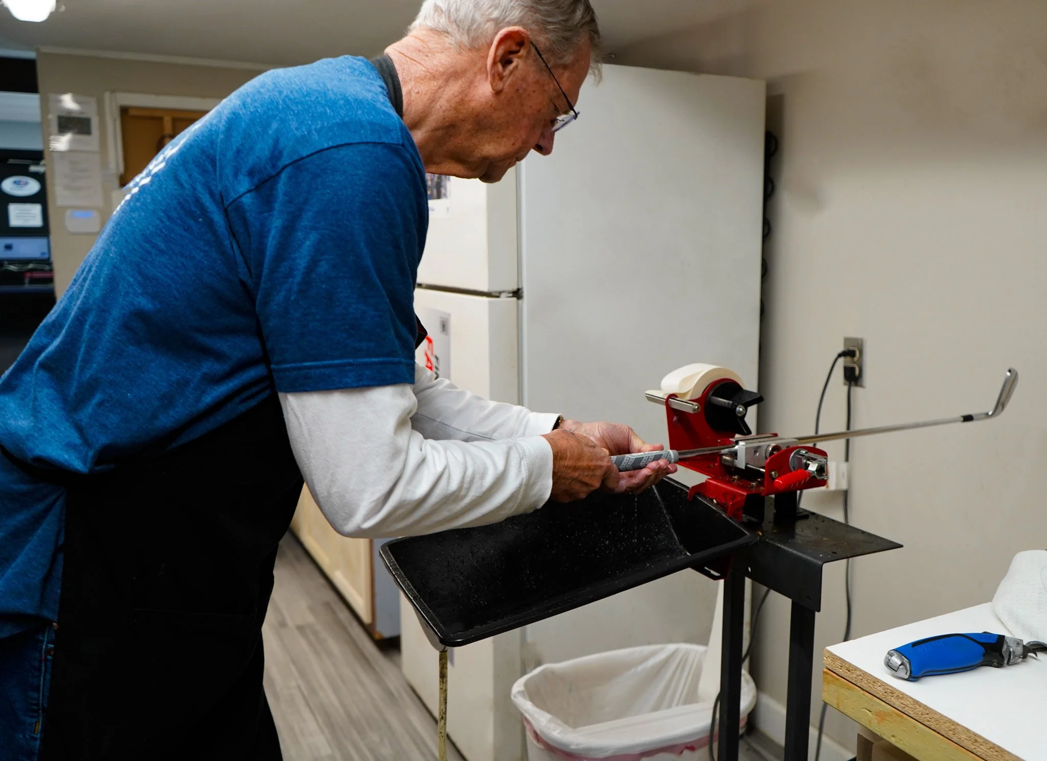 An elderly man with gray hair, wearing glasses, a blue shirt, and black apron, is operating a machine that is wrapping a metal rod or wire. The machine is red and white, and the man is focused on adjusting or working with it in a room with kitchen appliances and a white refrigerator in the background.