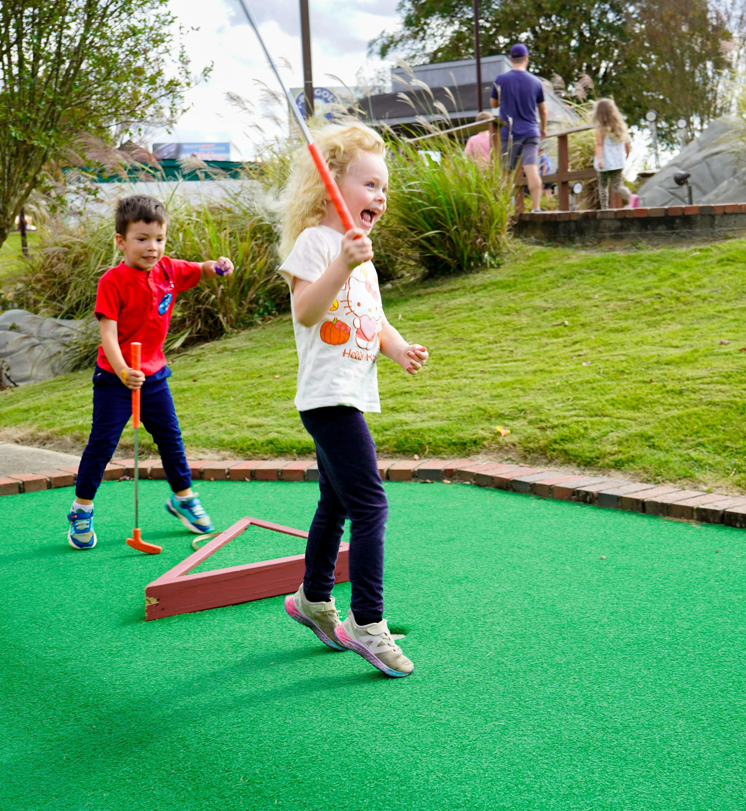 Children playing mini-golf on a green outdoor course with a girl swinging a golf club and a boy holding a golf ball behind her.