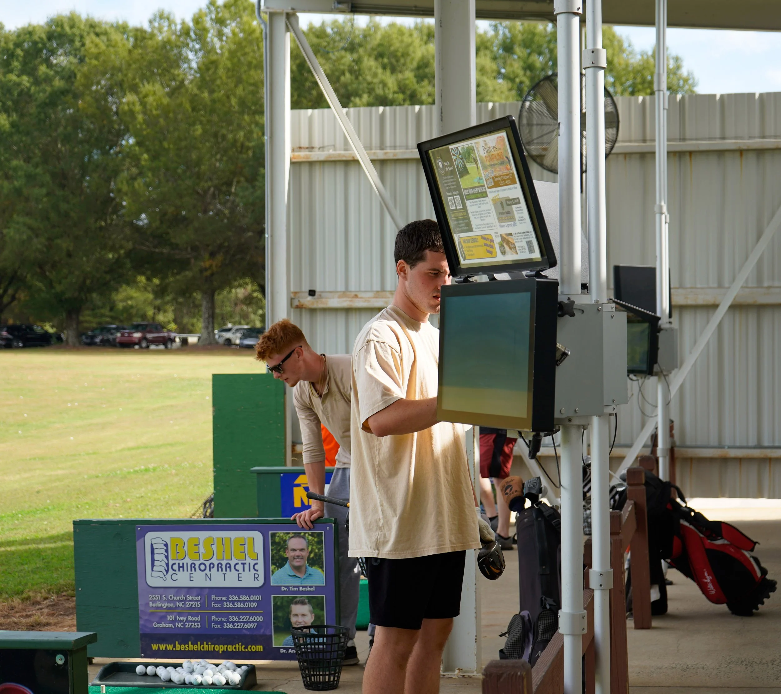 Two young men at a golf driving range, one is hitting golf balls with a club, and the other is looking at electronic scoreboards.