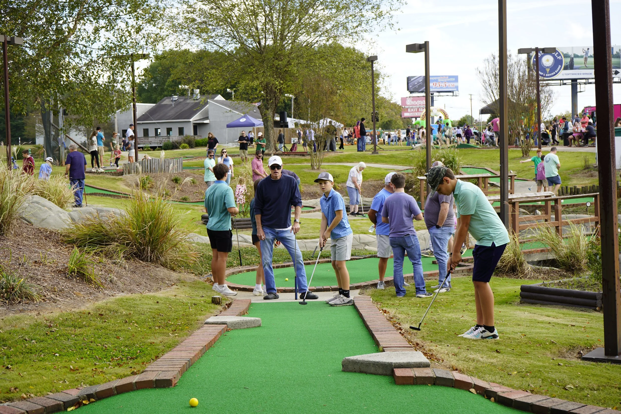 People playing mini golf on a bright, outdoor course with green artificial turf and small obstacles, surrounded by trees and landscaped bushes, with many visitors and family groups in the background.