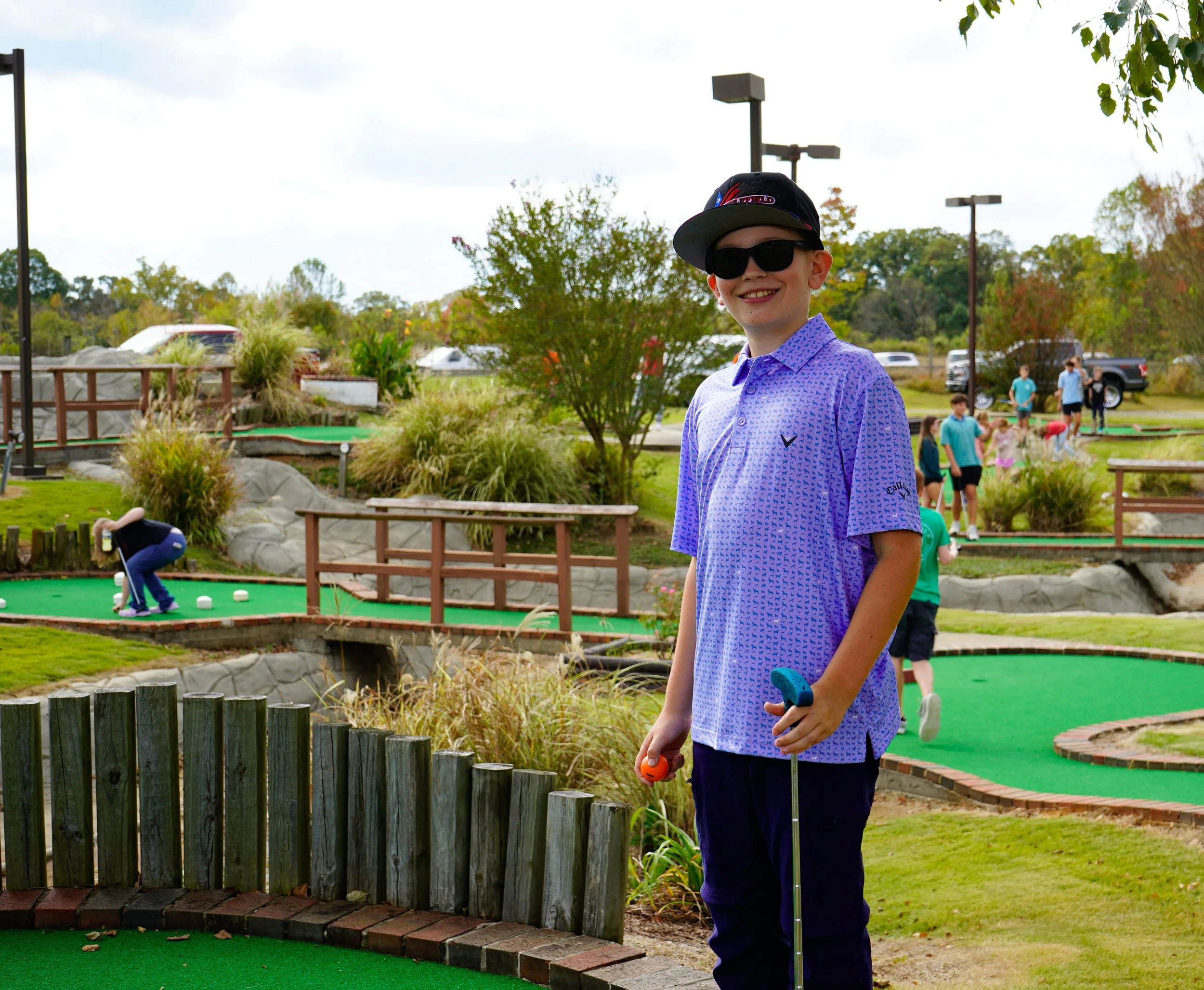 A young boy standing on a mini-golf course, wearing a purple polo shirt, sunglasses, a cap, and holding a mini-golf club. In the background, other children are playing mini-golf, and there are lush plants, rocks, and trees surrounding the course.