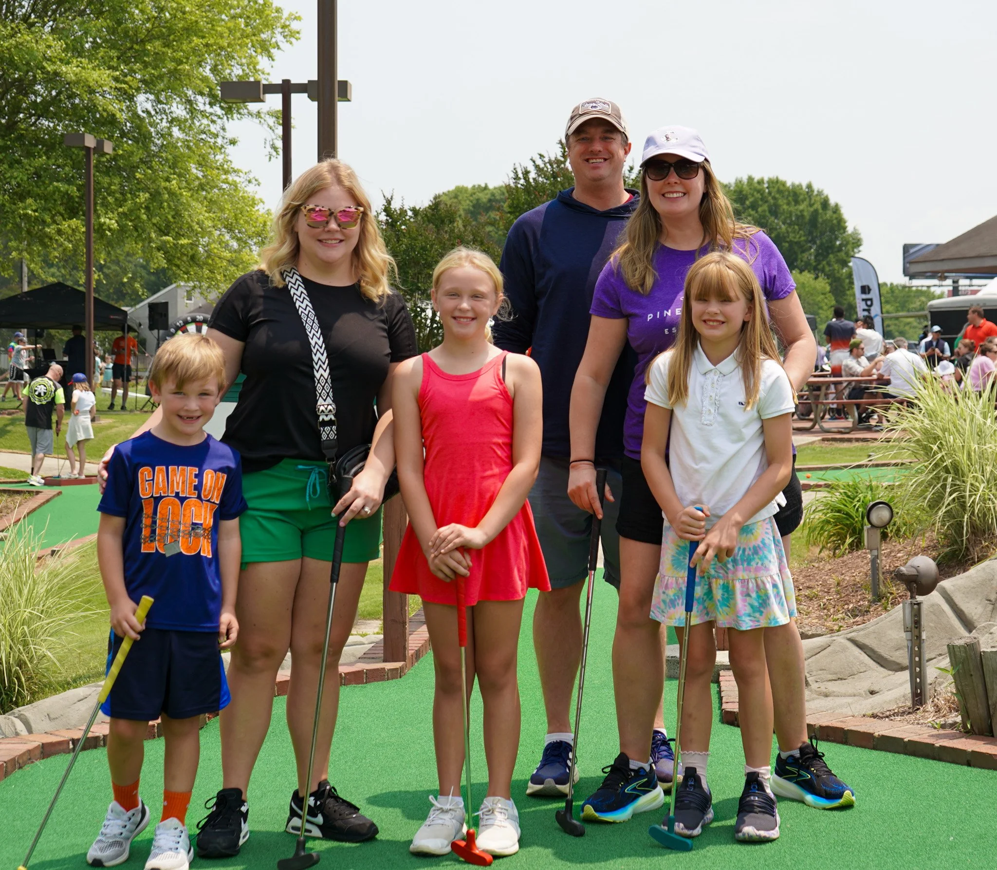 Family of six at a mini golf course, posing with golf clubs, smiling, outdoor setting, sunny day.