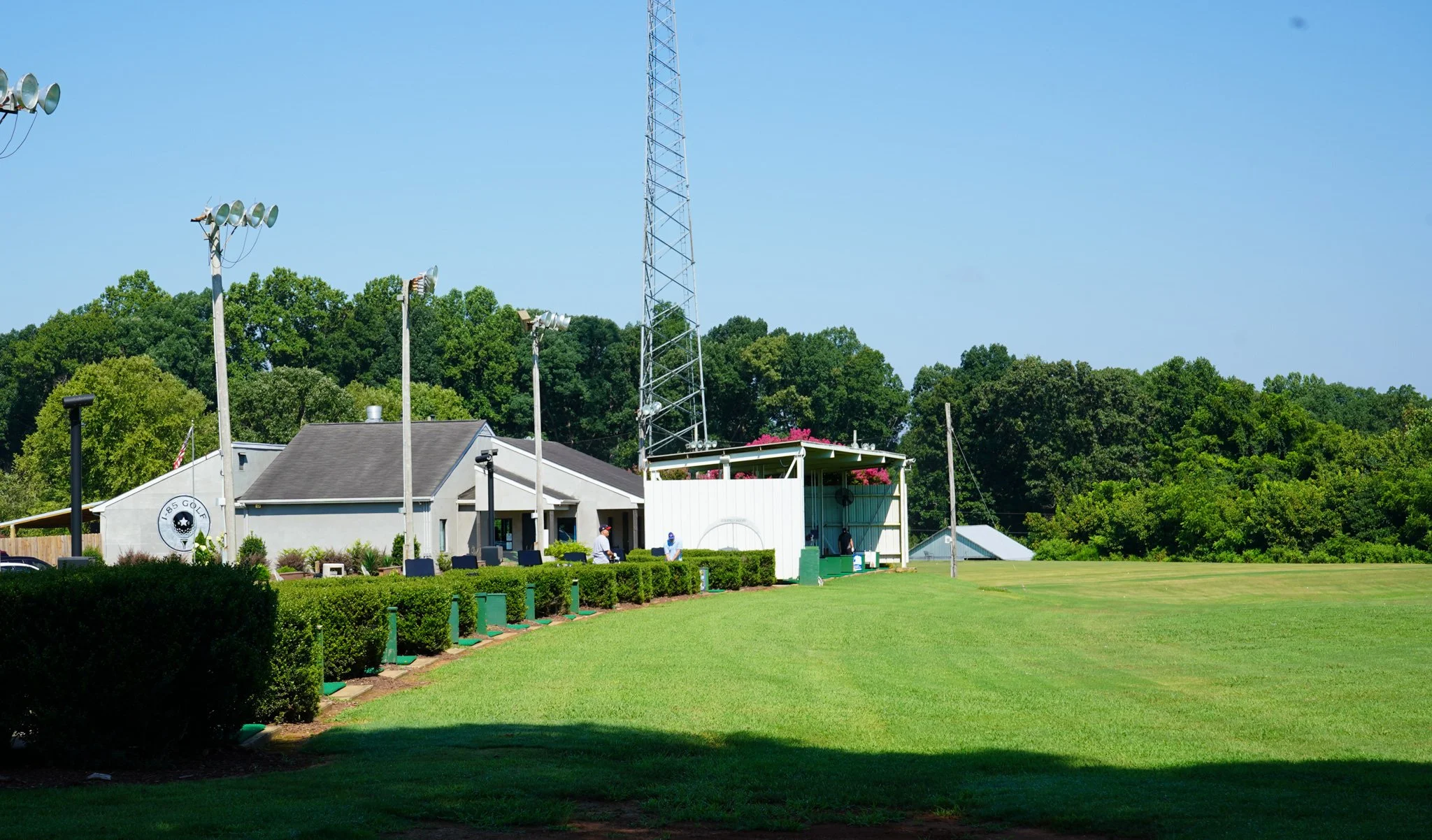 A golf driving range with a building, tall lights, and a large radio tower in the background. The area is surrounded by green trees and a well-maintained grassy field.