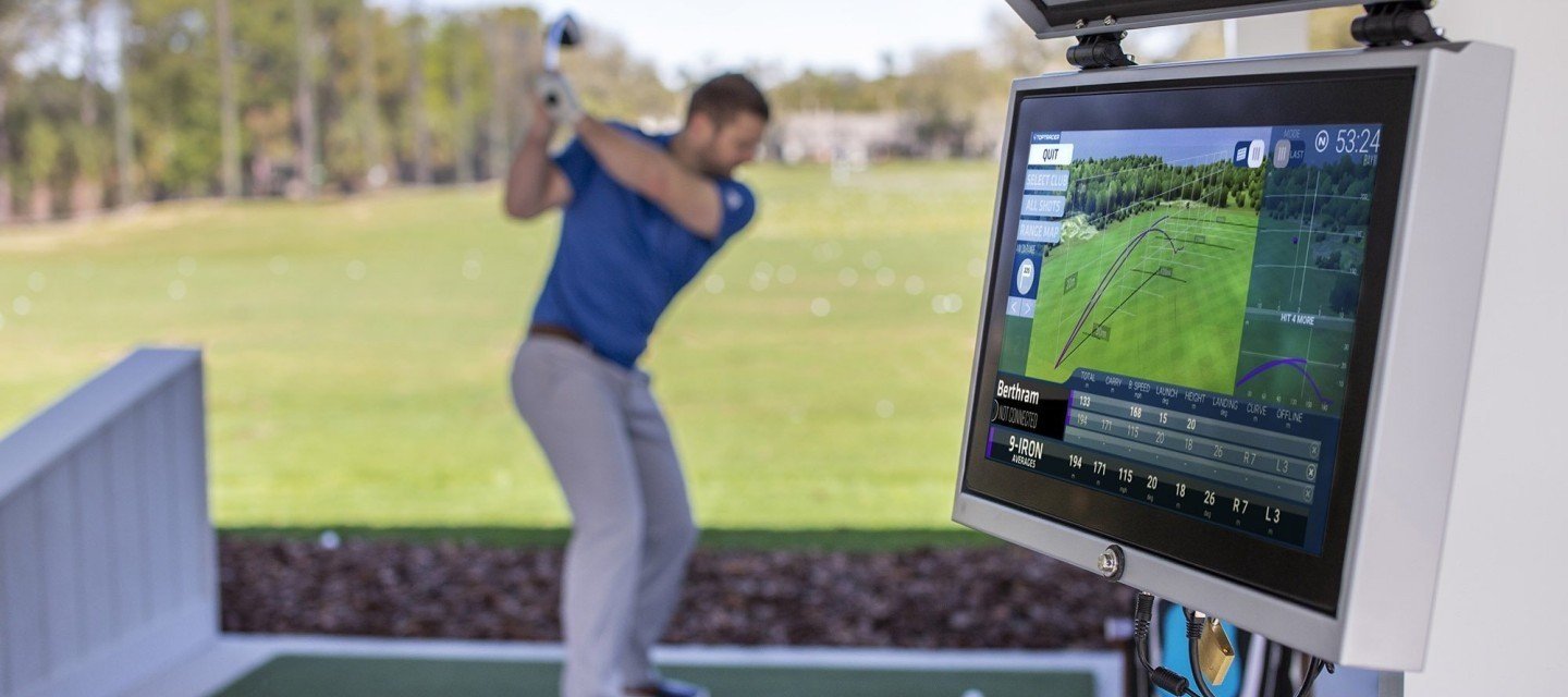 A man in a blue shirt practicing golf swings outdoors on a sunny day, with a digital golf simulator screen displaying swing analysis and trajectory data in the foreground.