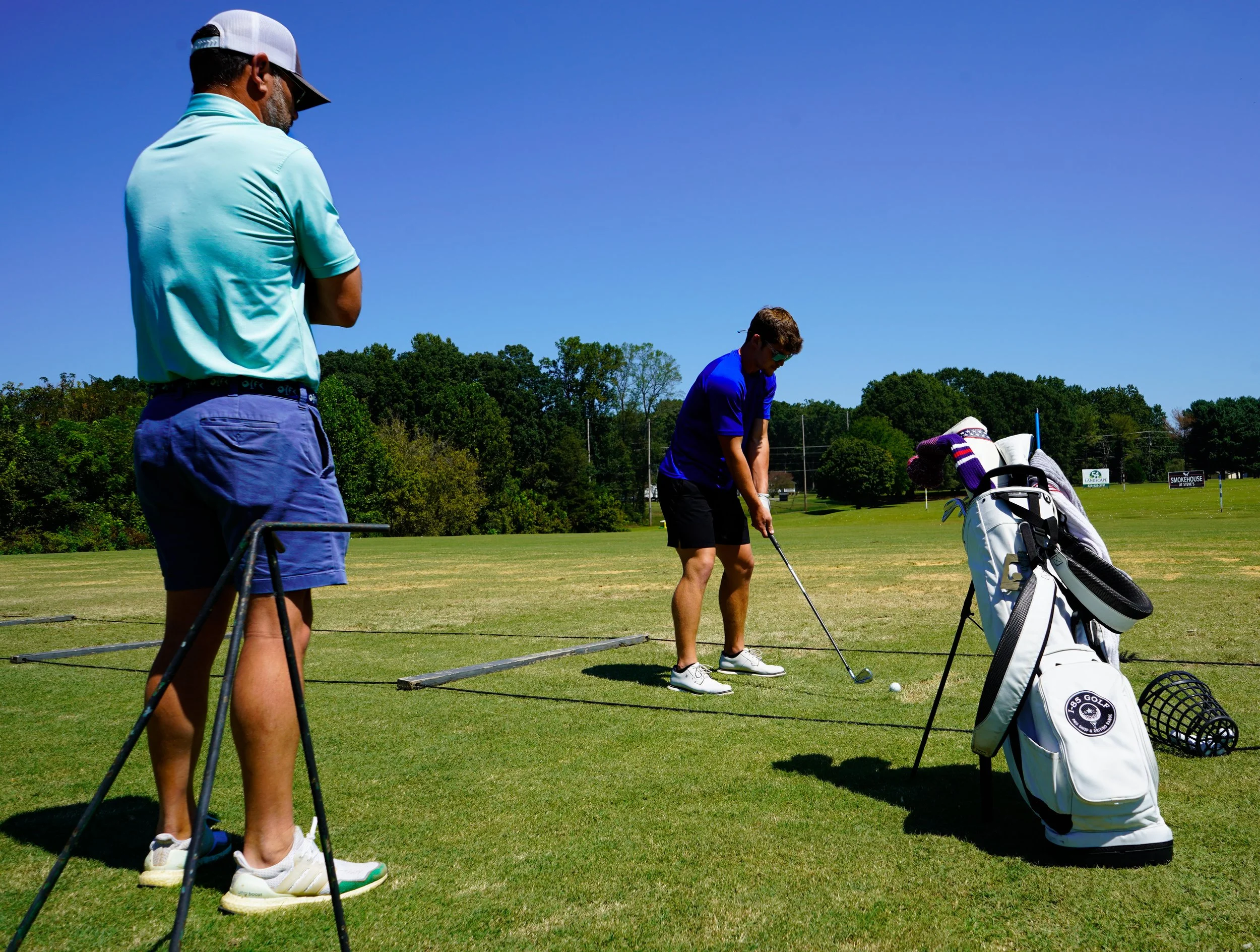 Two men on a golf driving range, one preparing to hit the ball while the other watches, with a golf bag and clubs nearby, under a clear blue sky.