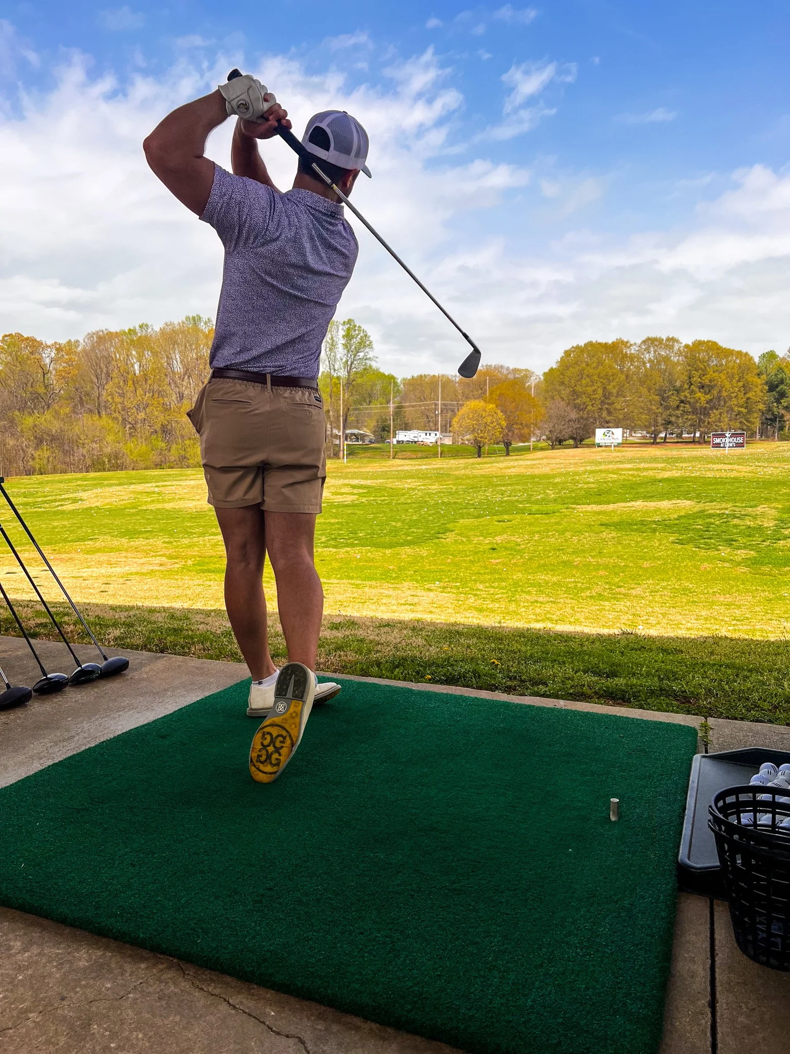 A man dressed in shorts, a polo shirt, and a cap is swinging a golf club on a golf course.