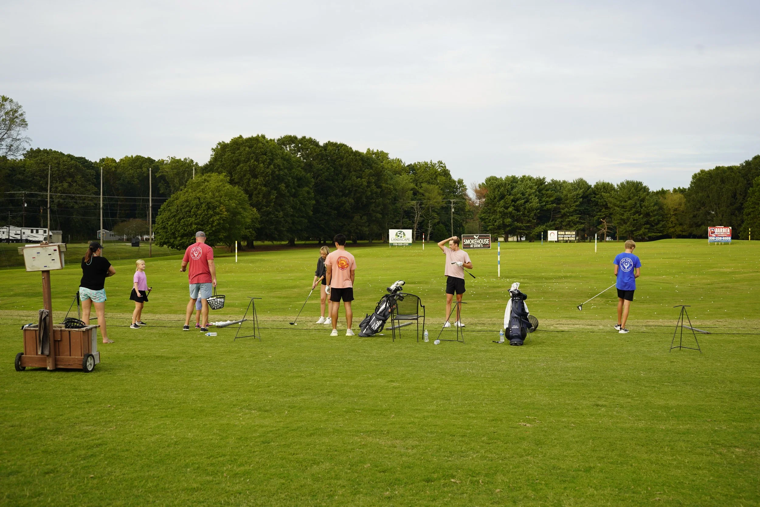 Group of people practicing golf on a driving range with trees and signs in the background.