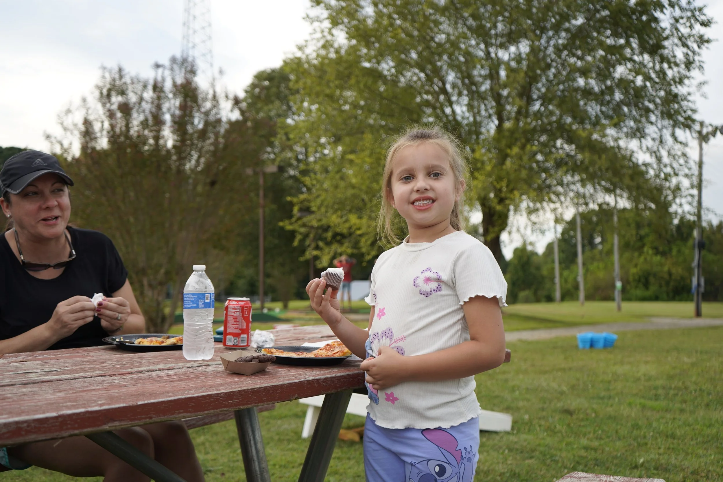 A young girl with blonde hair, wearing a white shirt with purple flowers and purple pajama pants featuring a cartoon character, stands outside at a picnic table holding a cupcake. An adult woman, sitting at the table, is smiling and holding her own cupcake. The table has pizza, a water bottle, a soda can, and small bowls. The background shows green trees and a cloudy sky.
