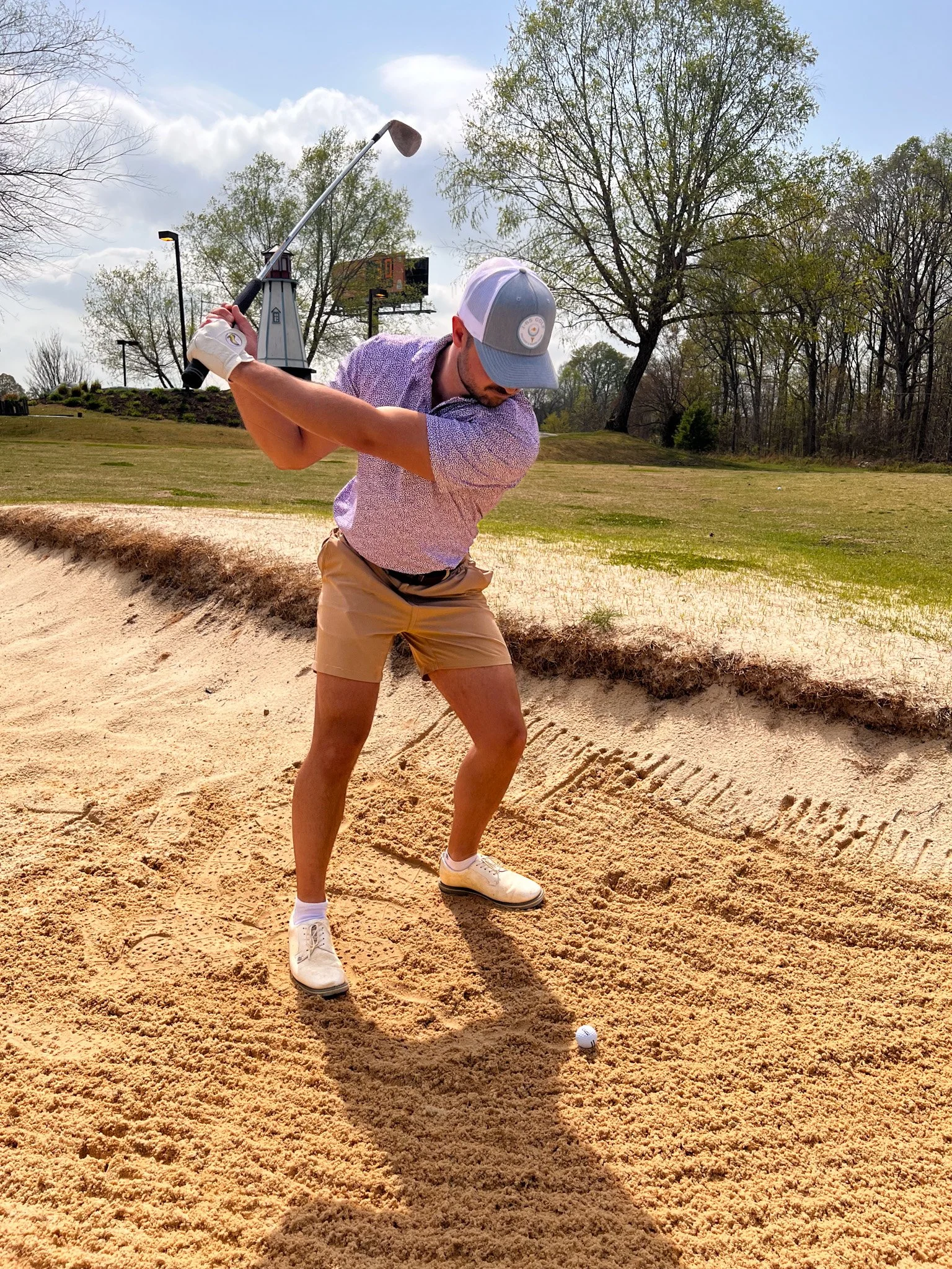 A man in a purple shirt, beige shorts, white shoes, and a cap is hitting a golf ball out of a sand bunker with a golf club, during daytime with partly cloudy sky.