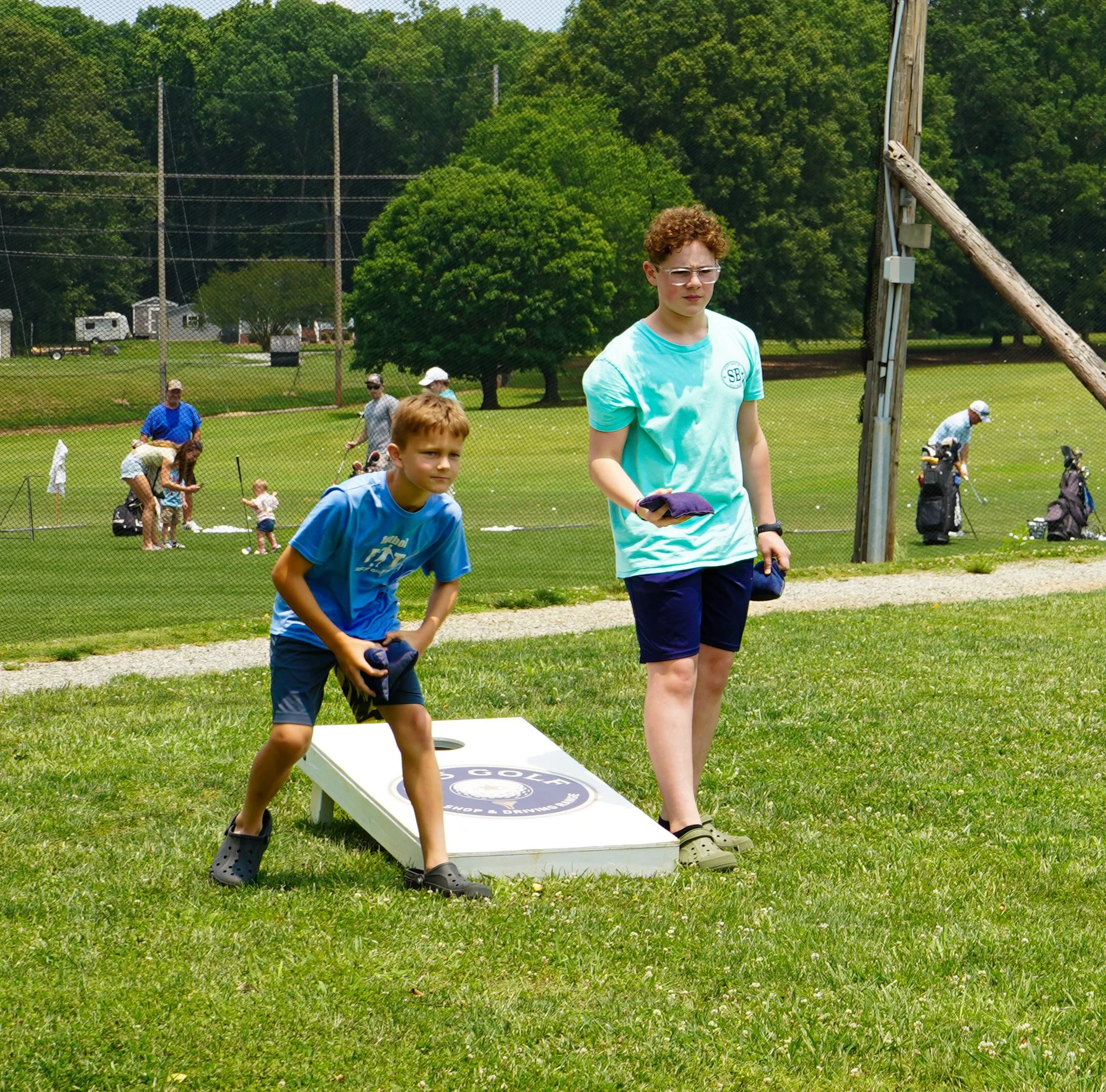 A young boy and a teenage girl playing cornhole outdoors on a grassy field during a sunny day, with a chain-link fence and people in the background.