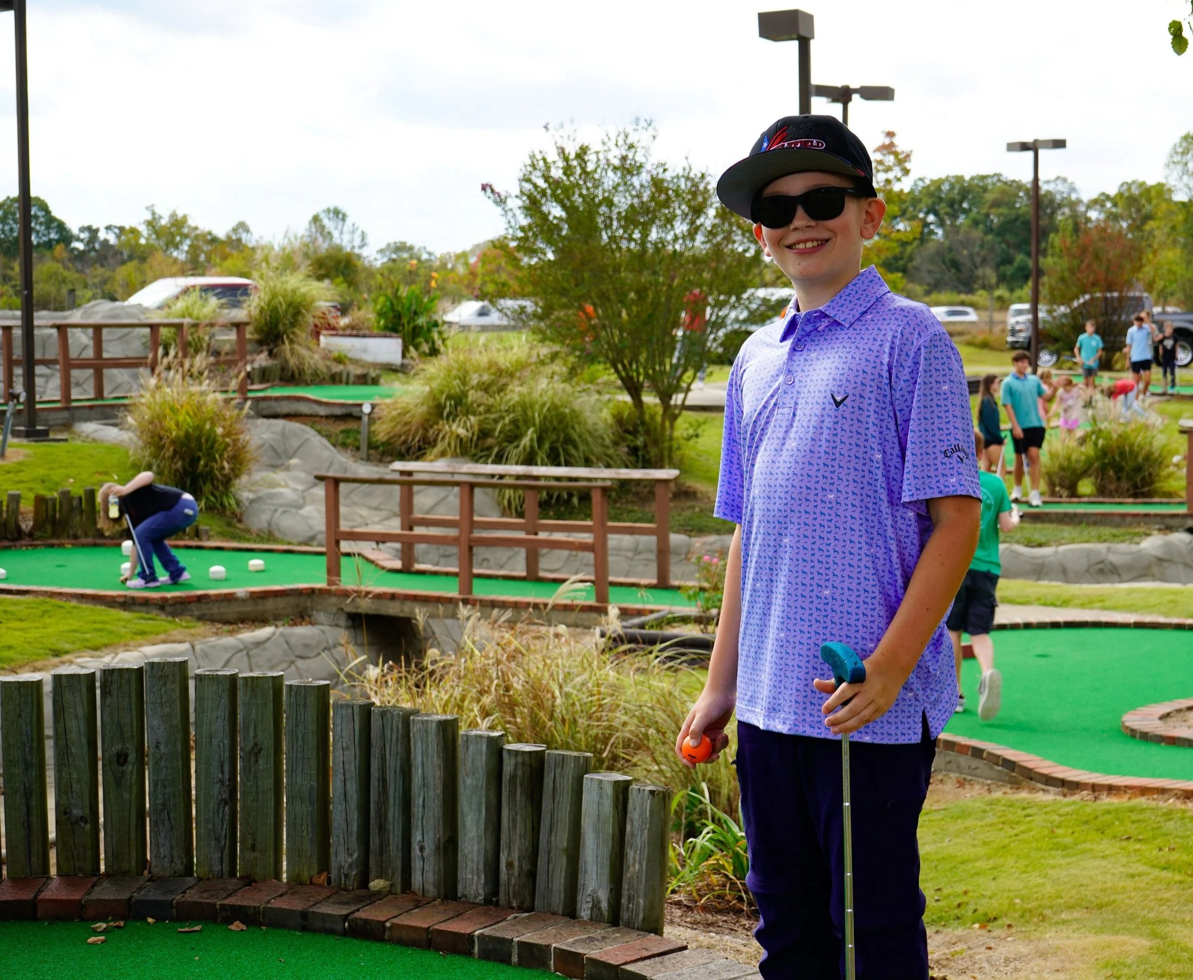 A smiling boy in sunglasses and a cap holding a mini golf club on a mini golf course with multiple players in the background.