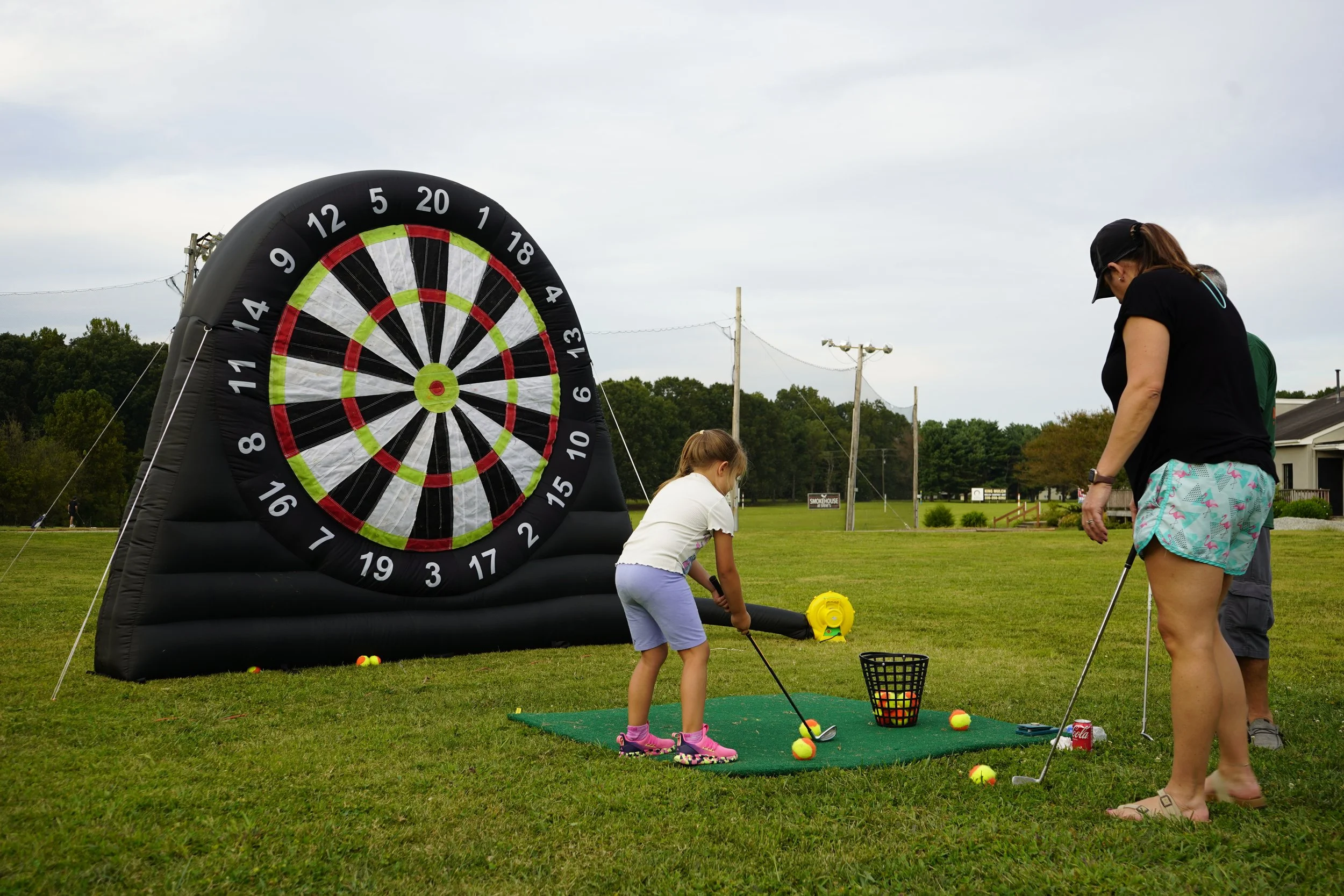 A girl practicing golf on a small mat with a woman supervising, next to a large inflatable dartboard in an open grassy area.
