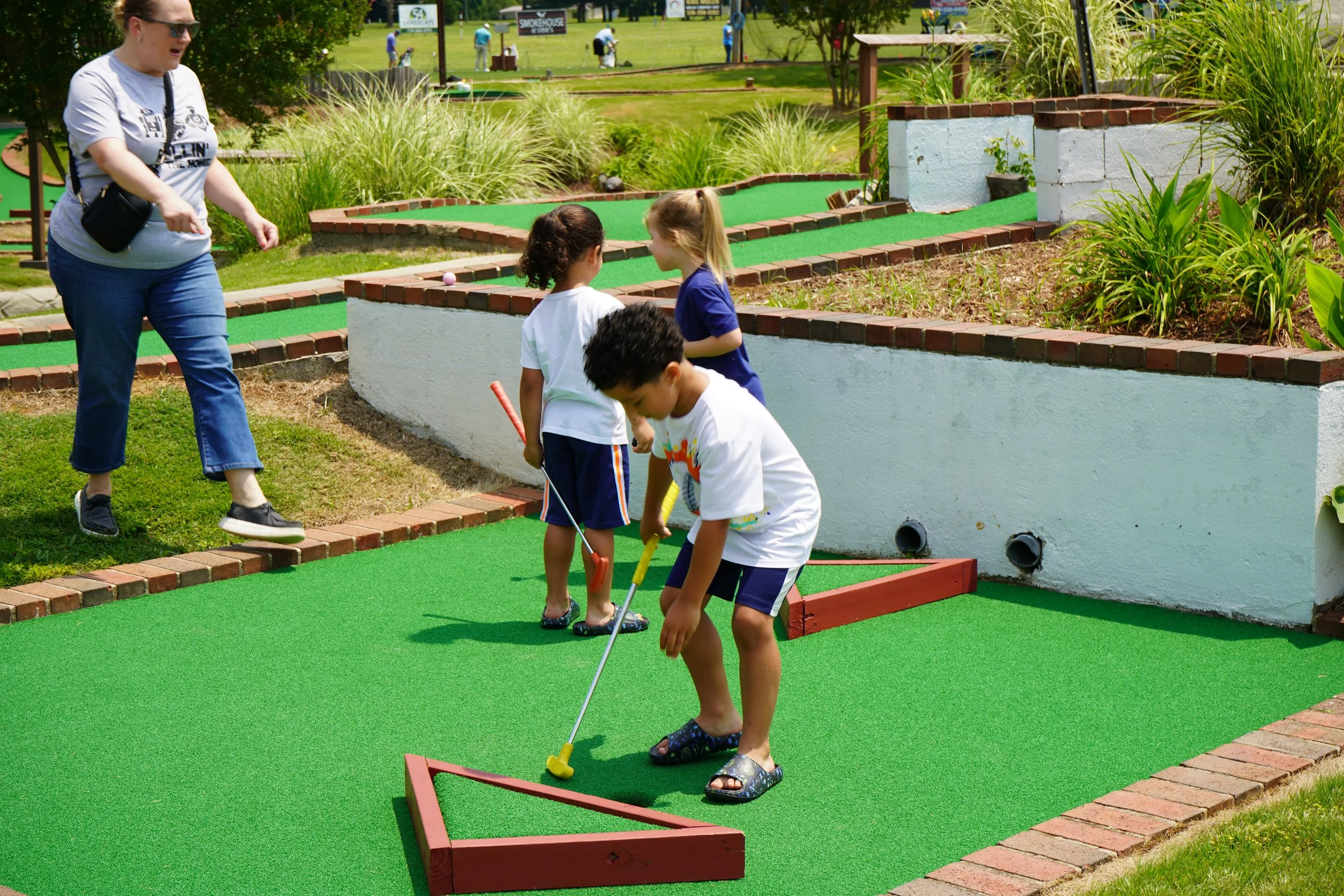 Children playing mini golf on a green course while an adult supervises, outdoor setting with plants and other people in the background.