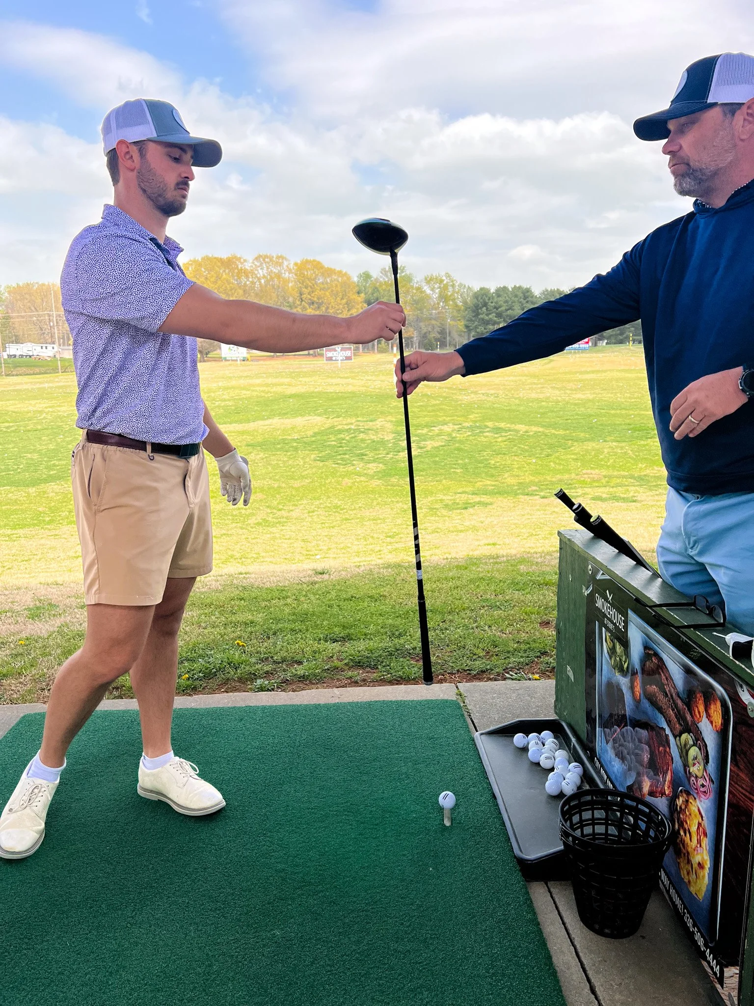 A young man wearing a cap, patterned shirt, tan shorts, and white shoes practicing golf at a driving range, assisted by a man in a dark shirt and cap, holding a golf club.