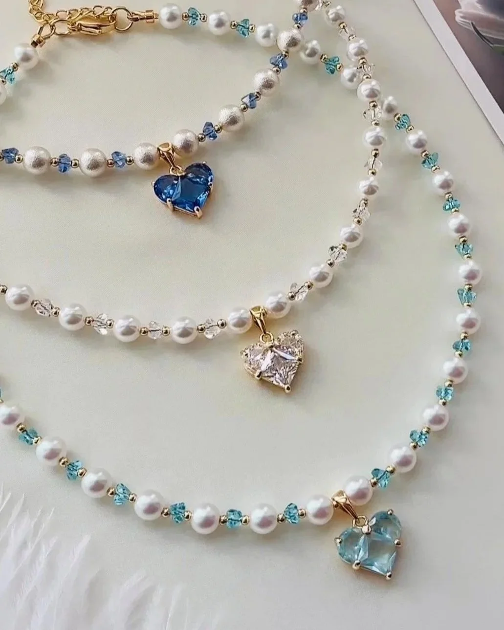 Three pearl necklaces with heart-shaped pendants in blue, clear, and light blue colors, laid out on a light-colored surface.