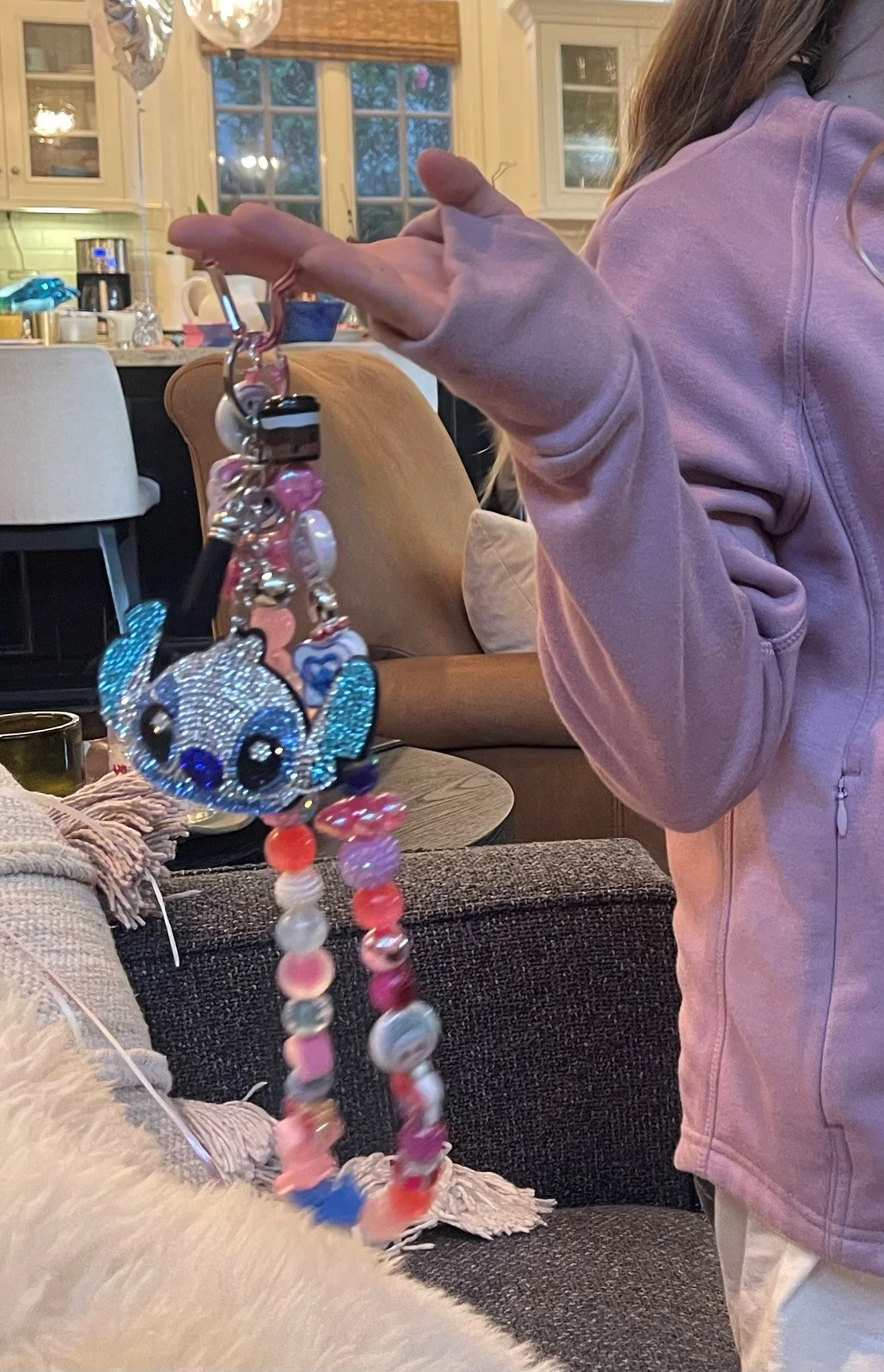 Person in pink jacket holding a keychain with a sparkly blue rhinestone fish and pink and white beaded strands, in a cozy living room setting.
