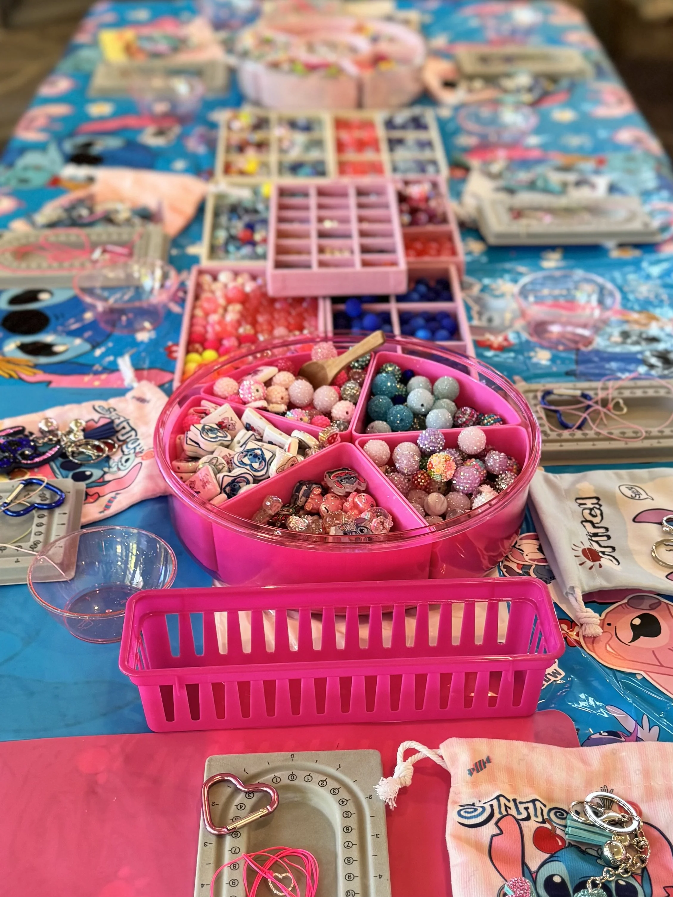 A table covered with a blue tablecloth featuring cartoon characters, displaying various jewelry-making supplies such as beads, charms, jewelry hooks, and threading materials in organized containers and baskets.