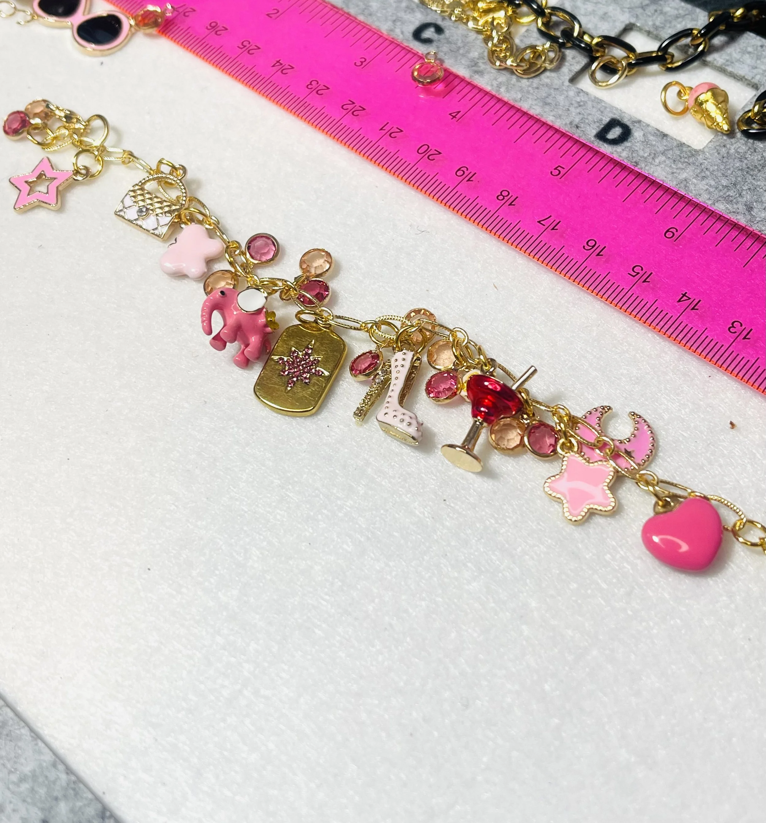 Pink charm bracelet with various charms including a star, moon, teddy bear, pink rectangle, high heel, heart, and cocktail glass, positioned near a pink ruler.