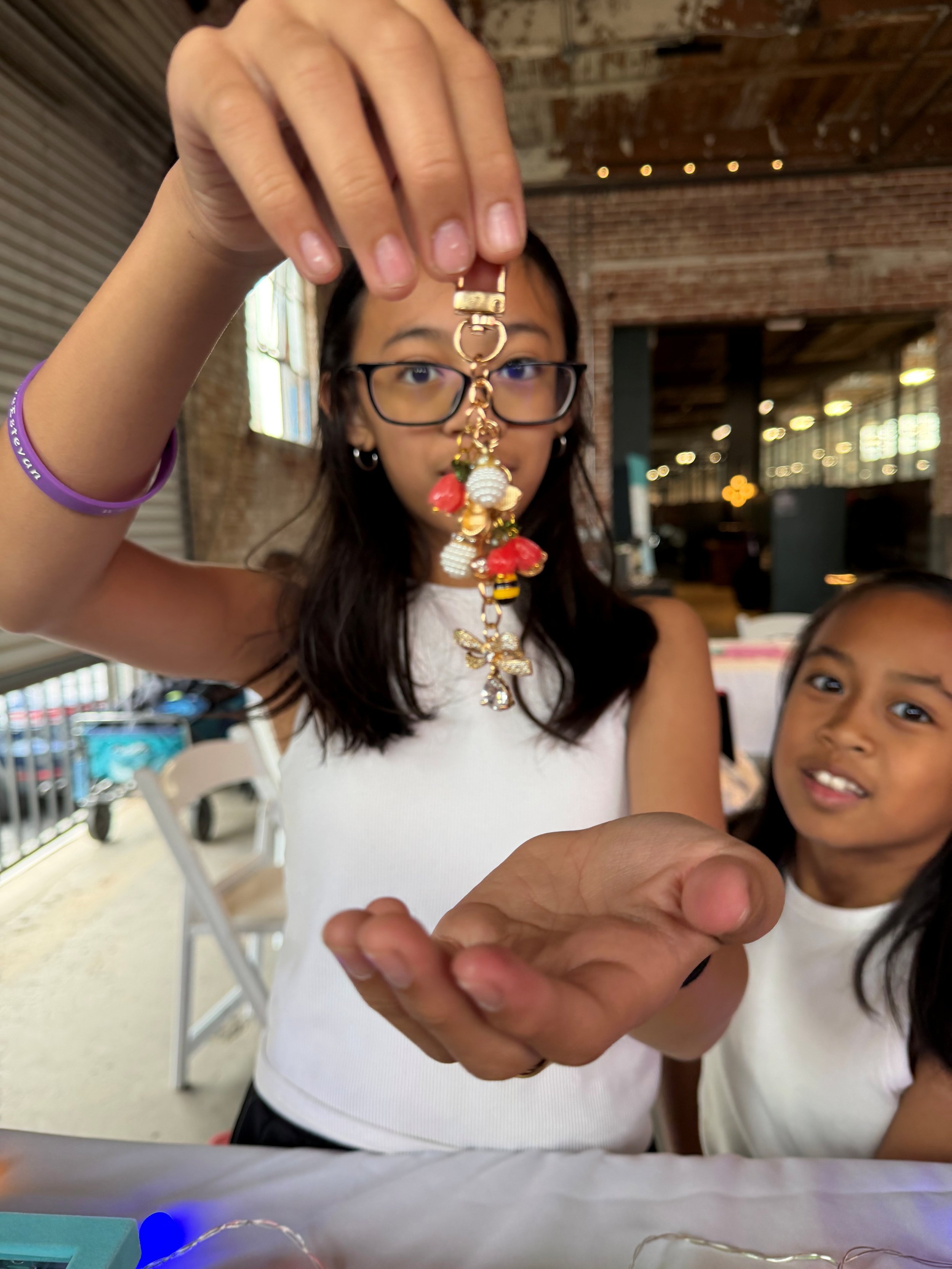 The image shows a girl with long dark hair and glasses holding up a gold-colored keychain with various charms, including a bee and white beads. Another girl with long dark hair is partially visible, looking at the camera. The background features a br