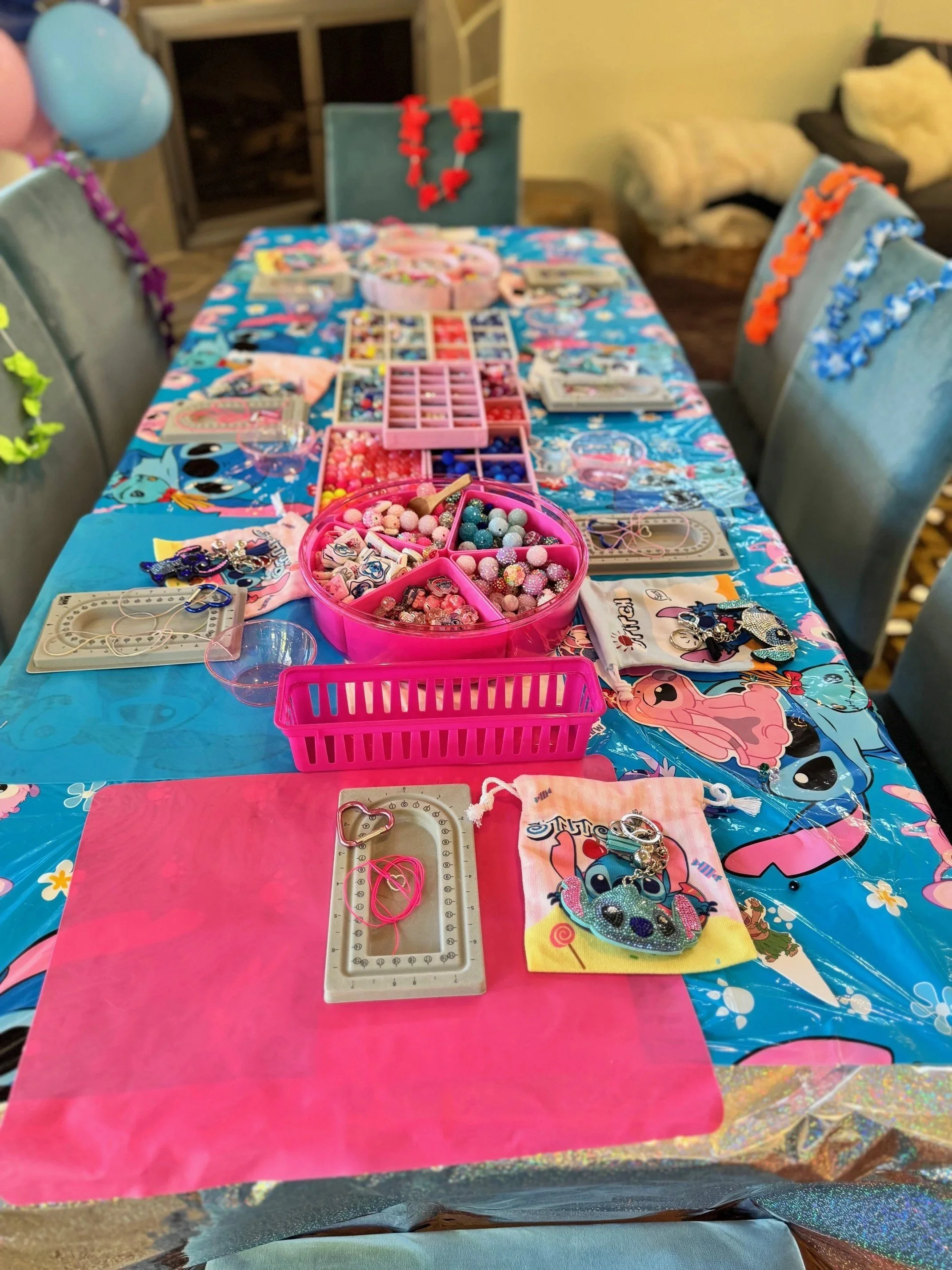 A decorated table with various buttons, beads, jewelry-making supplies, and craft materials. The table has a bright, colorful tablecloth featuring cartoon characters. There are chairs around the table decorated with colorful garlands. The background 
