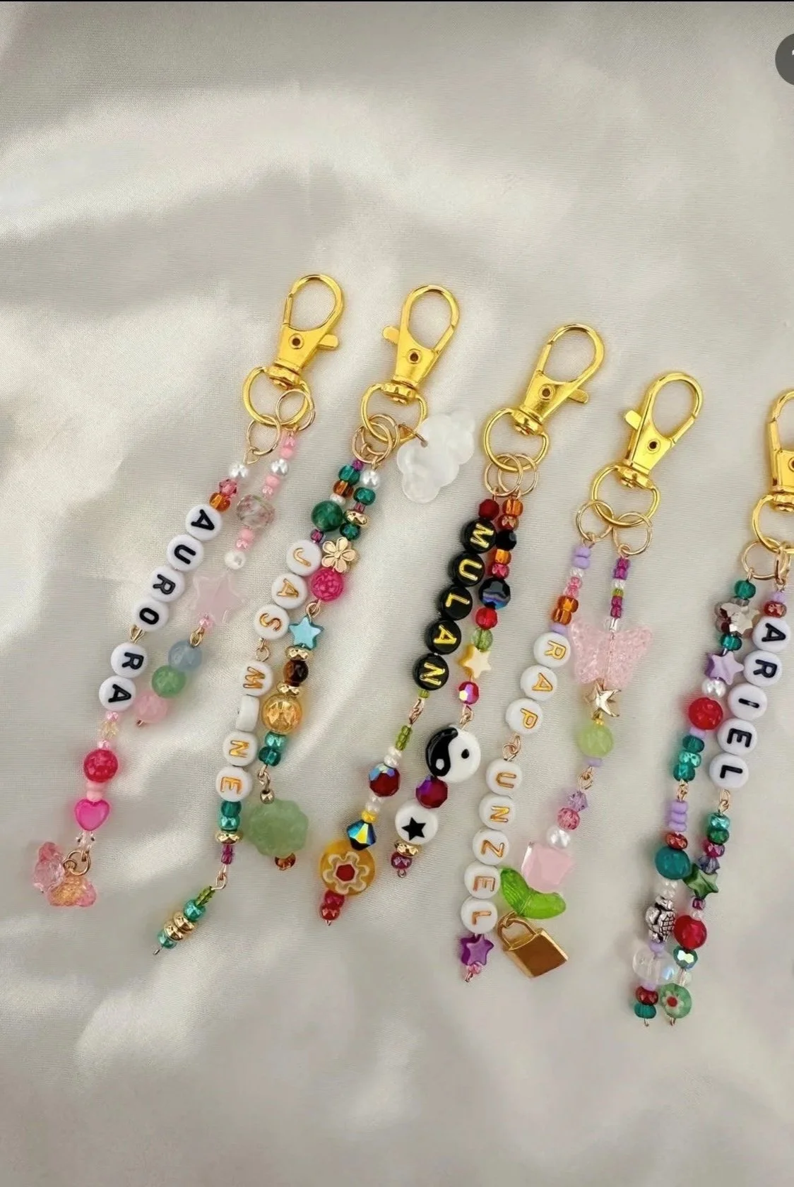 Several colorful beaded keychains with gold-tone clips, featuring various beads, charms, and letters spelling out names like Aurora, Savan, Autumn, and Ariel.