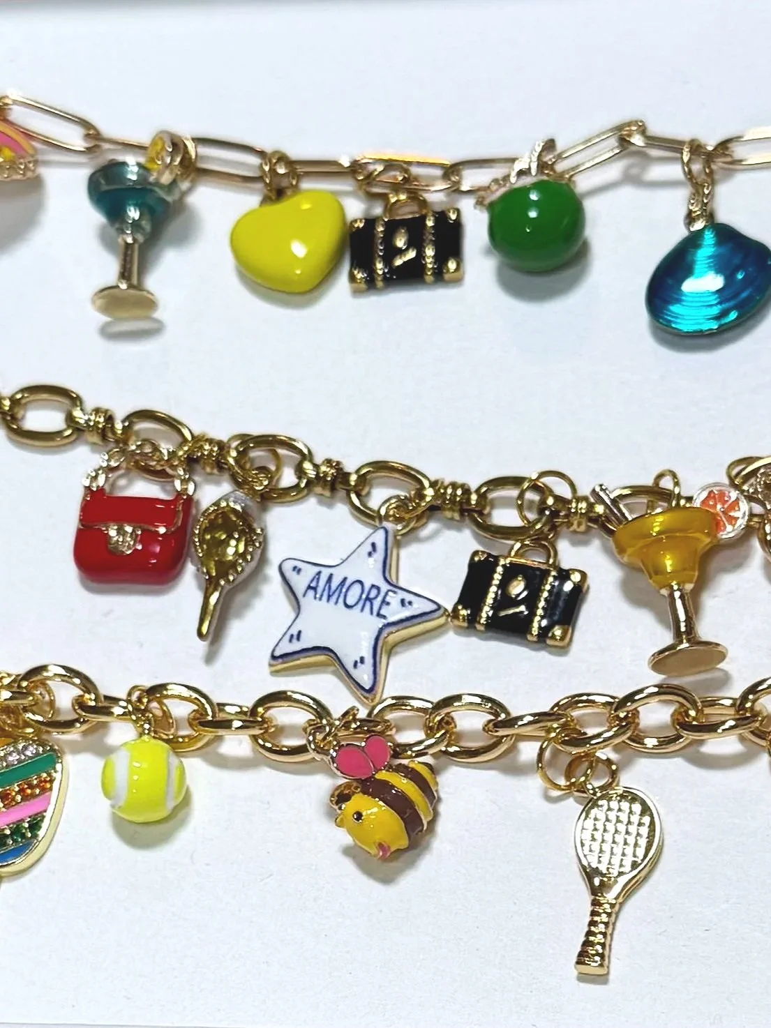 Three gold-colored charm bracelets with various colorful charms, including a star with 'AMORE' written on it, a tennis racket, a bee, a purse, a camera, a lollipop, a tennis ball, and a gift box.