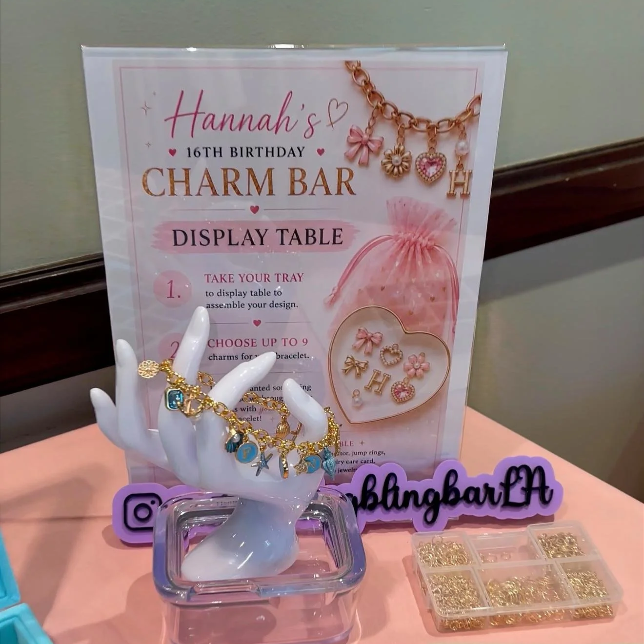 Display table with jewelry charms, including letter charms, flower charms, and heart charms, arranged on a tray and in plastic containers, with a sign promoting Hannah's charm bar for her 16th birthday, and decorative elements like a jewelry bracelet