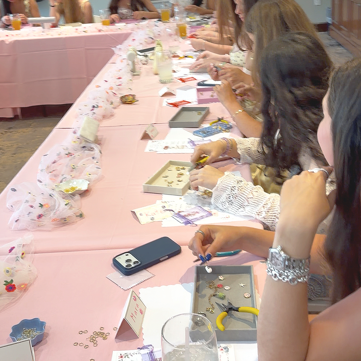 Group of women sitting at a long pink table, making jewelry with beads and charms during a craft event.
