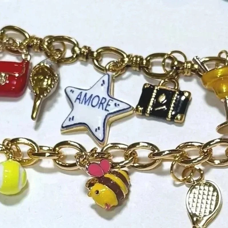 Gold charm bracelet with various colorful charms including a red backpack, starfish with 'AMORE' written on it, a camera, a yellow cocktail glass, a pink and yellow bee, a tennis ball, and other charms.