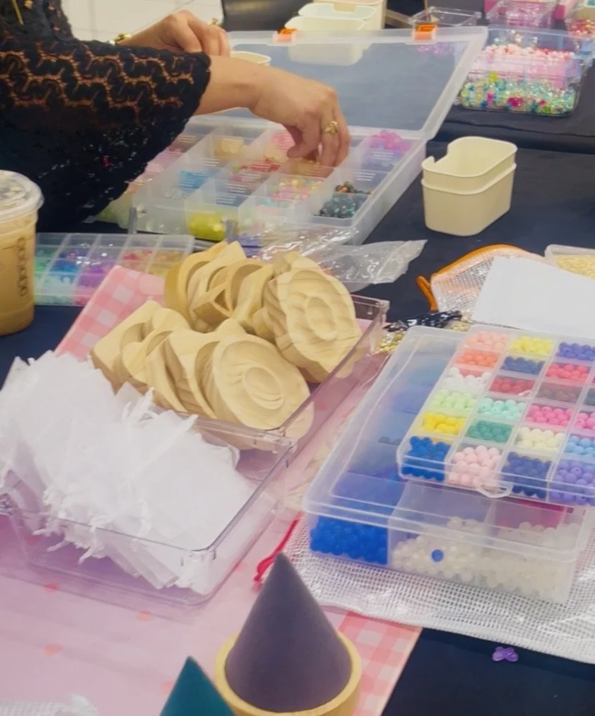 A person working with colorful beads, wooden craft pieces, and various craft supplies on a table covered with a pink checkered tablecloth. The table includes organized bead containers, a cone-shaped craft item, and other crafting tools.
