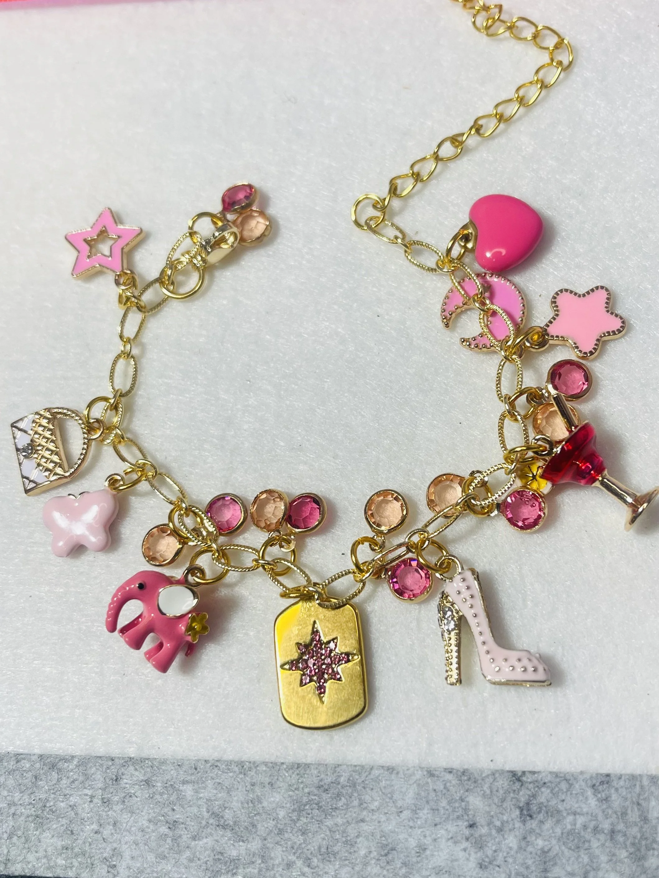 Gold charm bracelet with pink, peach, and white charms including a pink elephant, a pink and white high heel shoe, a gold dog tag with a pink star, a pink heart, a pink star, and pink, orange, and red gemstone accents on a white background. Feminine 