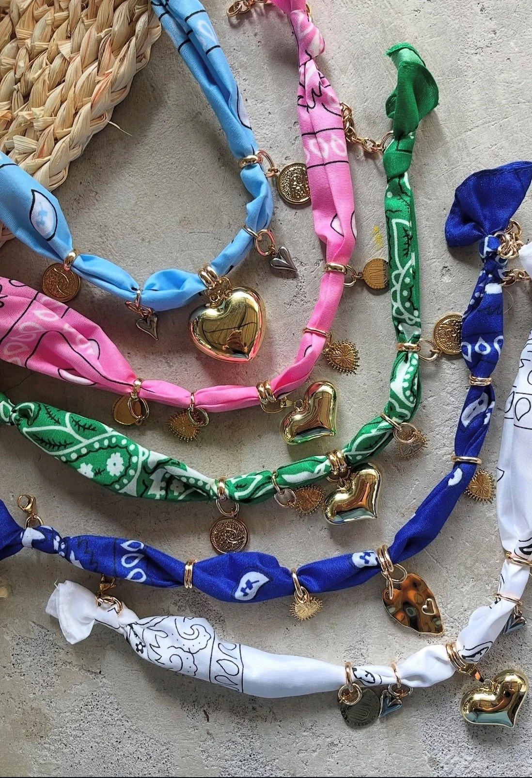 Colorful bandana necklaces with gold charms and heart-shaped pendants on a textured surface.