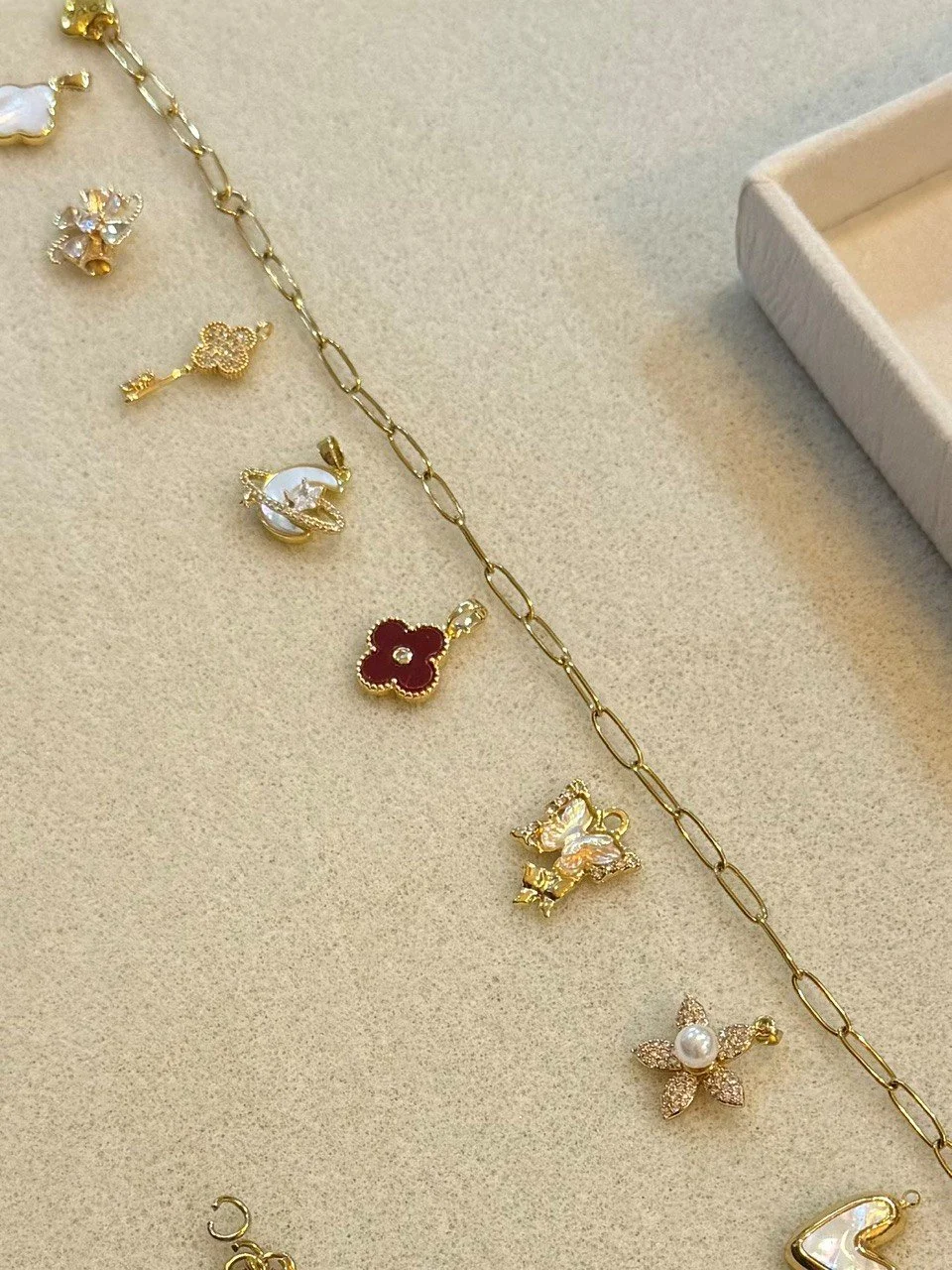 Collection of gold and silver jewelry charms, including a flower, a unicorn, a star, and other decorative pendants, spread out on a beige surface with an empty jewelry box.