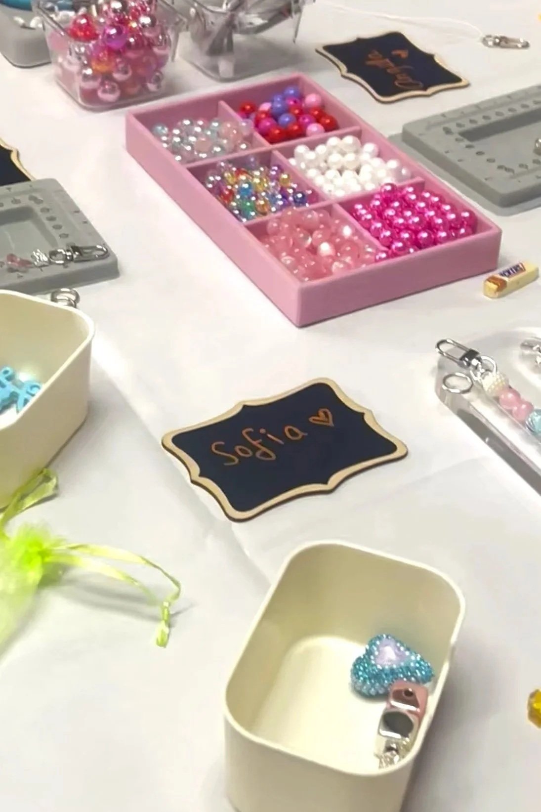Table display with various colorful beads and charms for jewelry making, including a pink bead box, small containers of beads, and a chalkboard sign that says 'Sofia' with a heart.