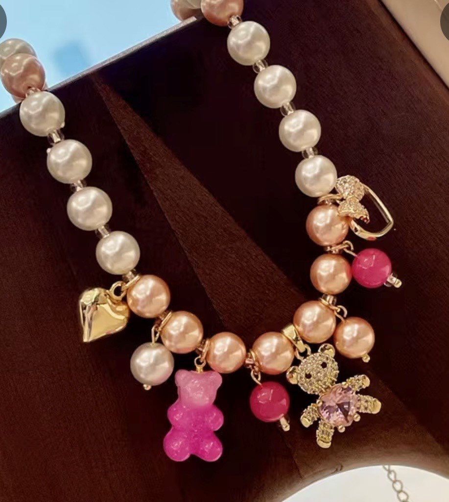 A pearl necklace with colorful charms, including a pink teddy bear, a bear adorned with rhinestones, and a gold heart, displayed on a brown wooden surface.