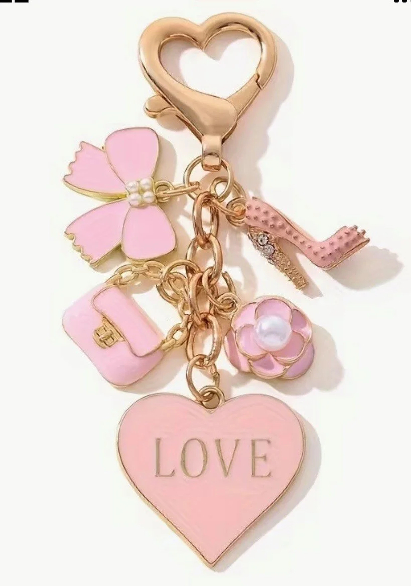 Pink and gold keychain with charms including a heart-shaped locket with the word 'LOVE', a butterfly, a purse, a flower with a pearl center, a letter 'L', and a small pink heart-shaped clasp.