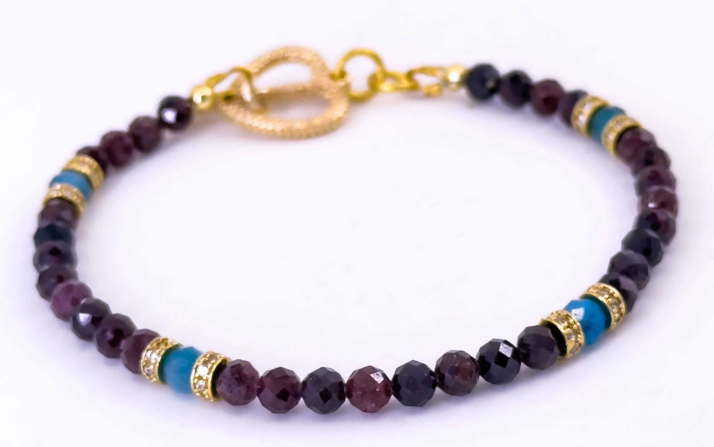 Beaded bracelet with dark purple, black, and maroon faceted beads, accented with gold and turquoise spacer beads, featuring a gold toggle clasp.
