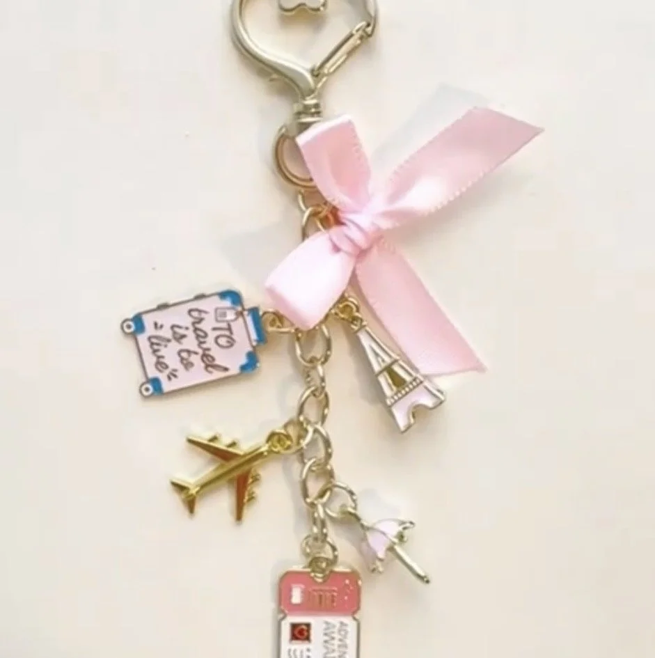 A simple yet elegant bag charm with multiple charms, including a pink ribbon bow, an Eiffel Tower, a small airplane, a miniature Instagram icon, a sign that says 'To travel is to live,' and a tiny manicure polish bottle.