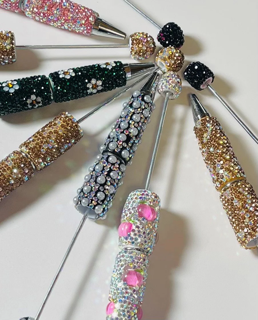 A close-up of decorative pens covered with various rhinestones and beads, arranged in a circular pattern on a white surface.