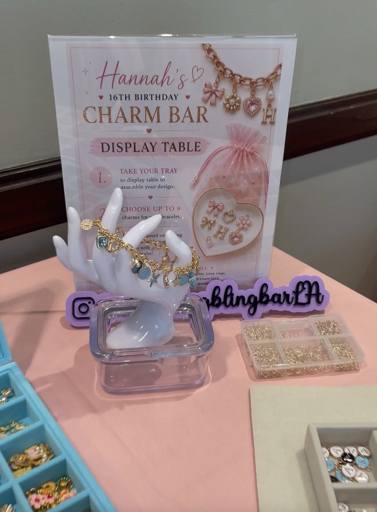 Display table with jewelry charms, bracelets, and decorative items, featuring a sign for Hannah's 16th birthday charm bar with instructions for creating a bracelet, and a jewelry hand model showcasing charms.
