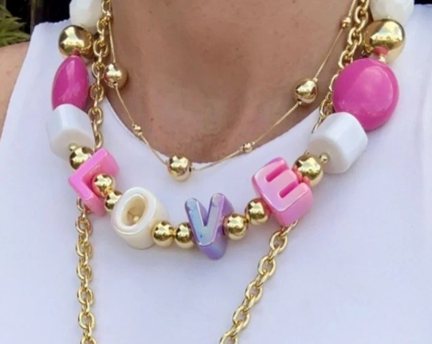 Close-up of a person's neck and chest wearing layered necklaces, one with beads spelling the word 'LOVE' in colorful letters, along with pink and white beads, and gold chains.