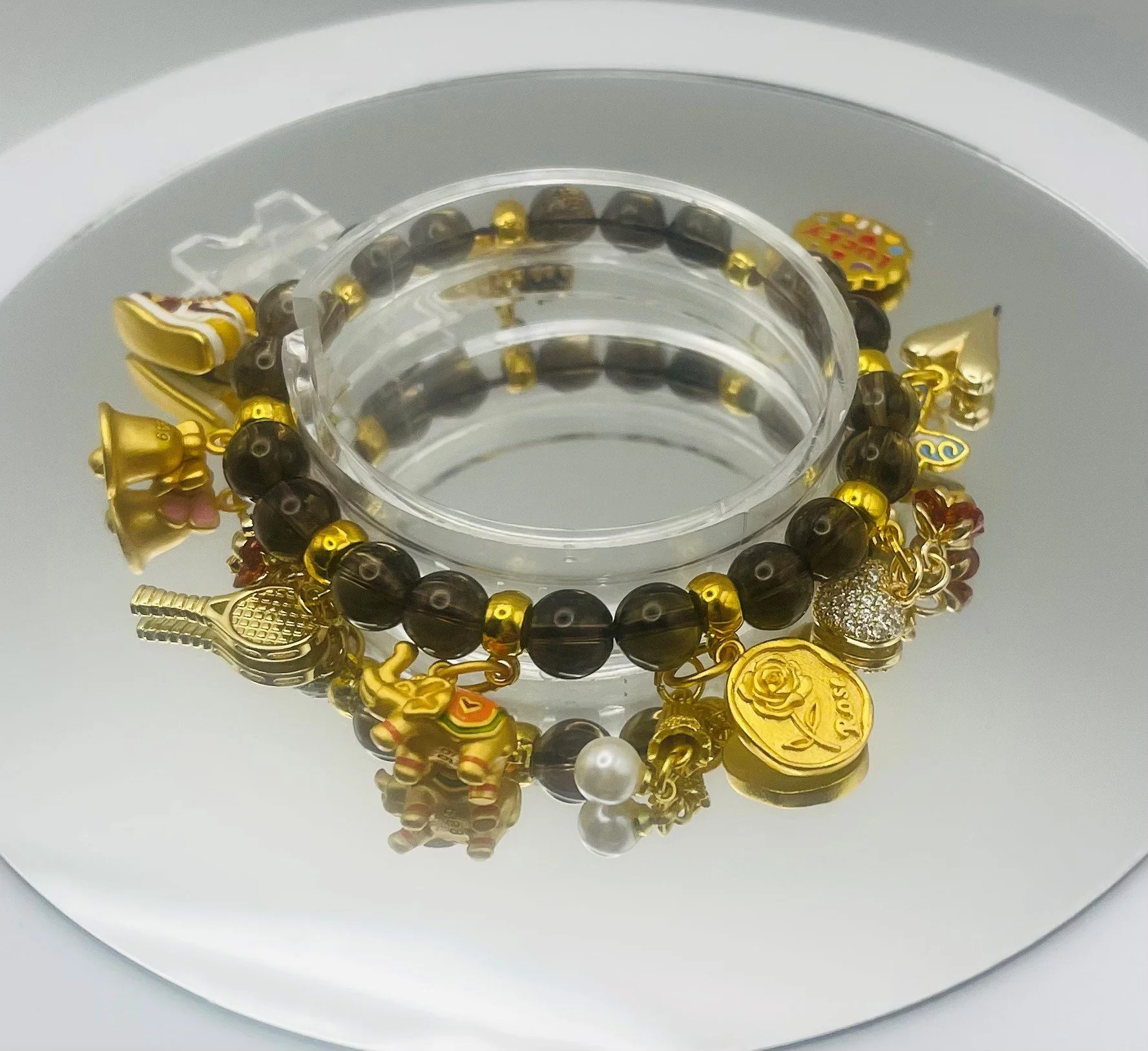 A circular glass container with a beaded bracelet, featuring black and gold beads, and gold charms including a rose, a heart, and a pineapple, is reflected on a mirror surface.