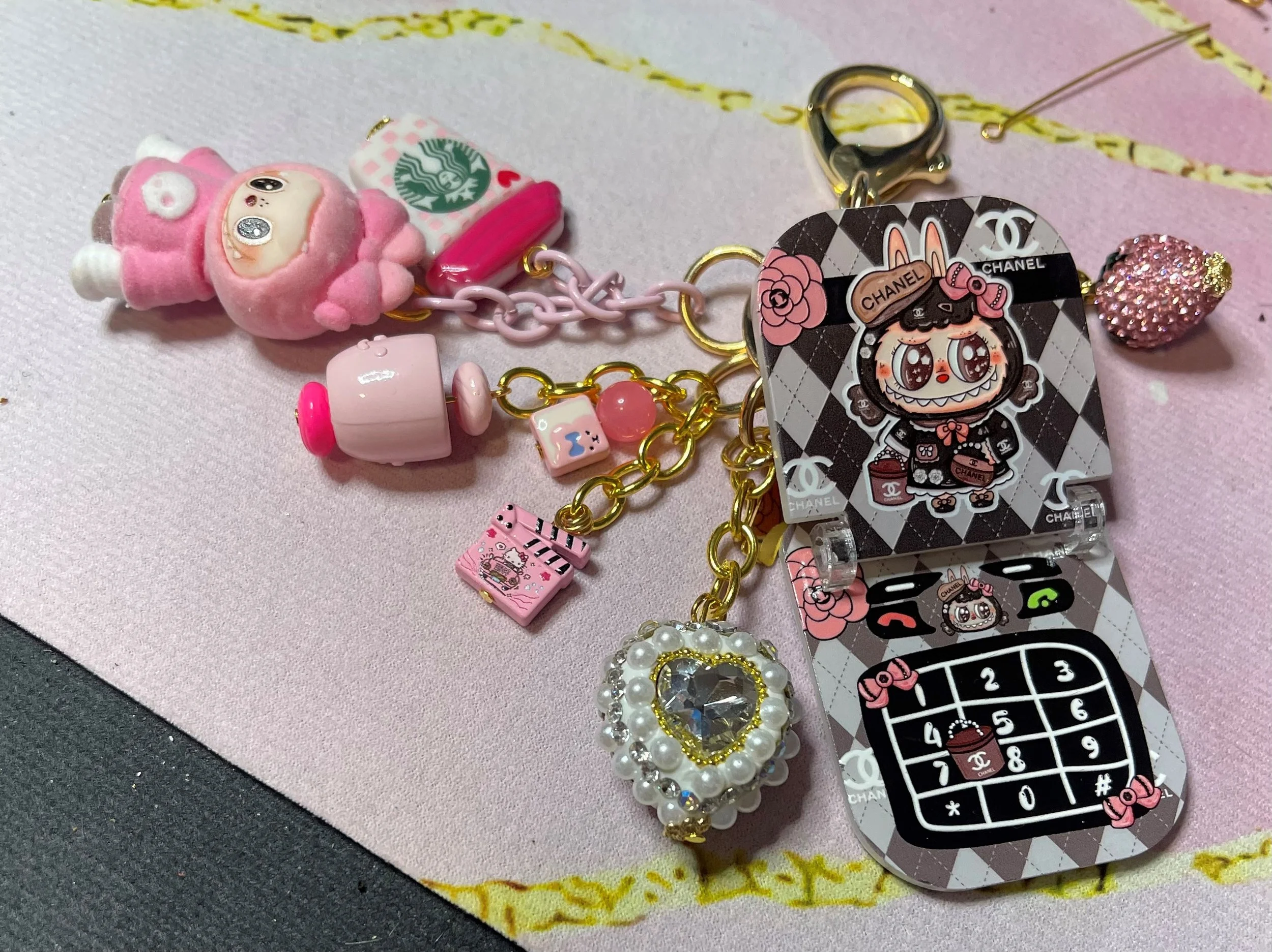 A labubu collection of colorful keychains and charms, including a pink sheep, Starbucks mug, a chanel-themed cartoon character, jeweled heart, and a decorative heart with pearls and stones, all lying on a pink surface.