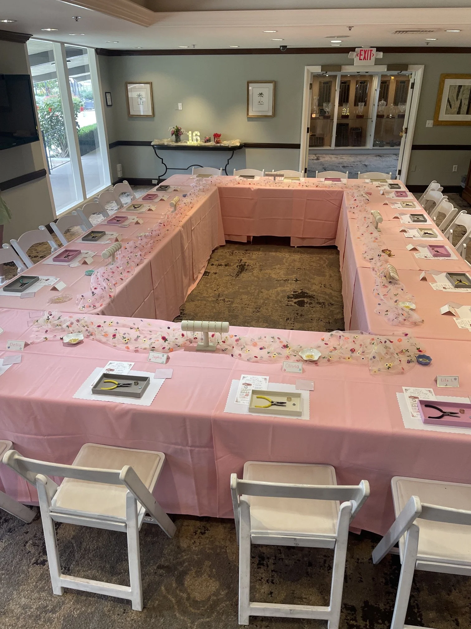 A U-shaped table set up for a celebration with pink tablecloths, decorated with a pink and floral runner, small plates, and place cards. There are white chairs around the table. In the background, there is a hallway with framed pictures and a side ta